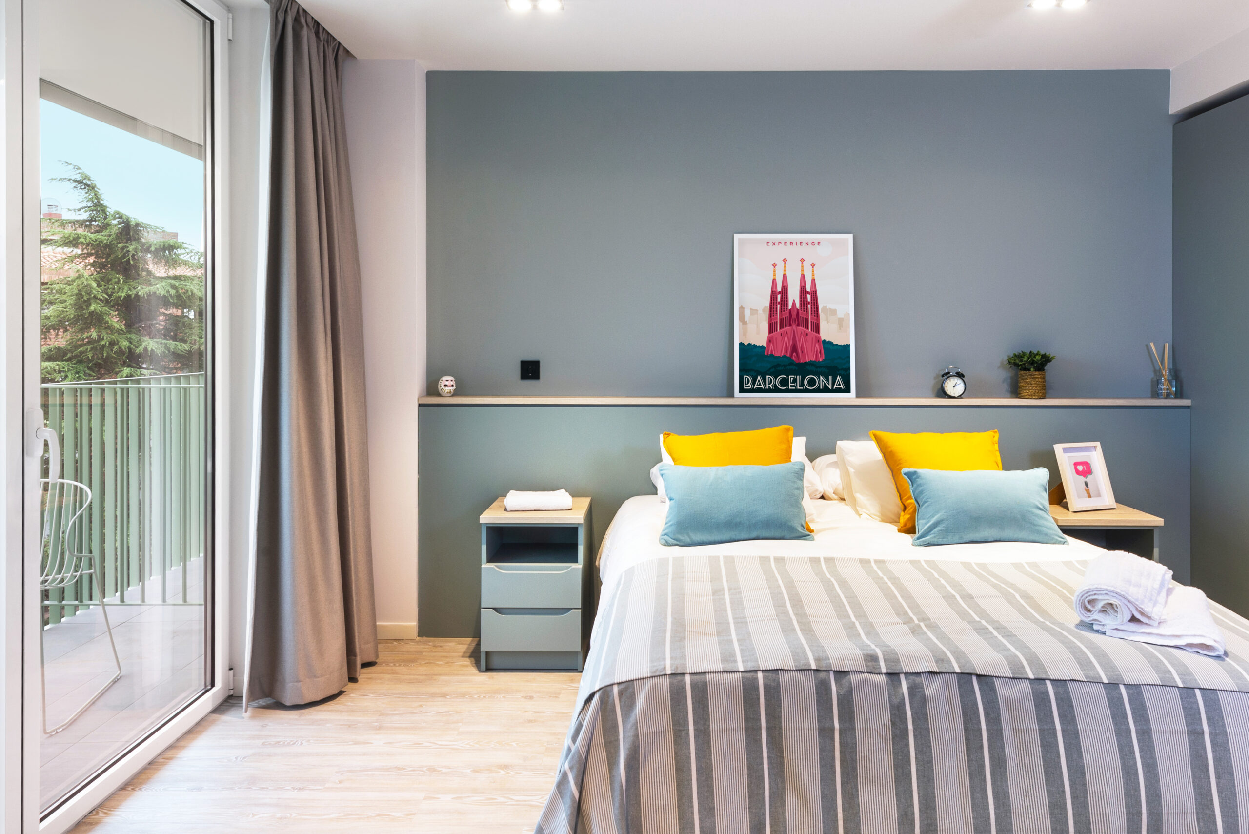 Modern bedroom with a large bed featuring striped bedding and colorful pillows. Above the bed is a Barcelona-themed artwork. Natural light streams in from a sliding glass door, leading to a balcony overlooking the trees of Bruce Street. Minimalist decor completes the space.