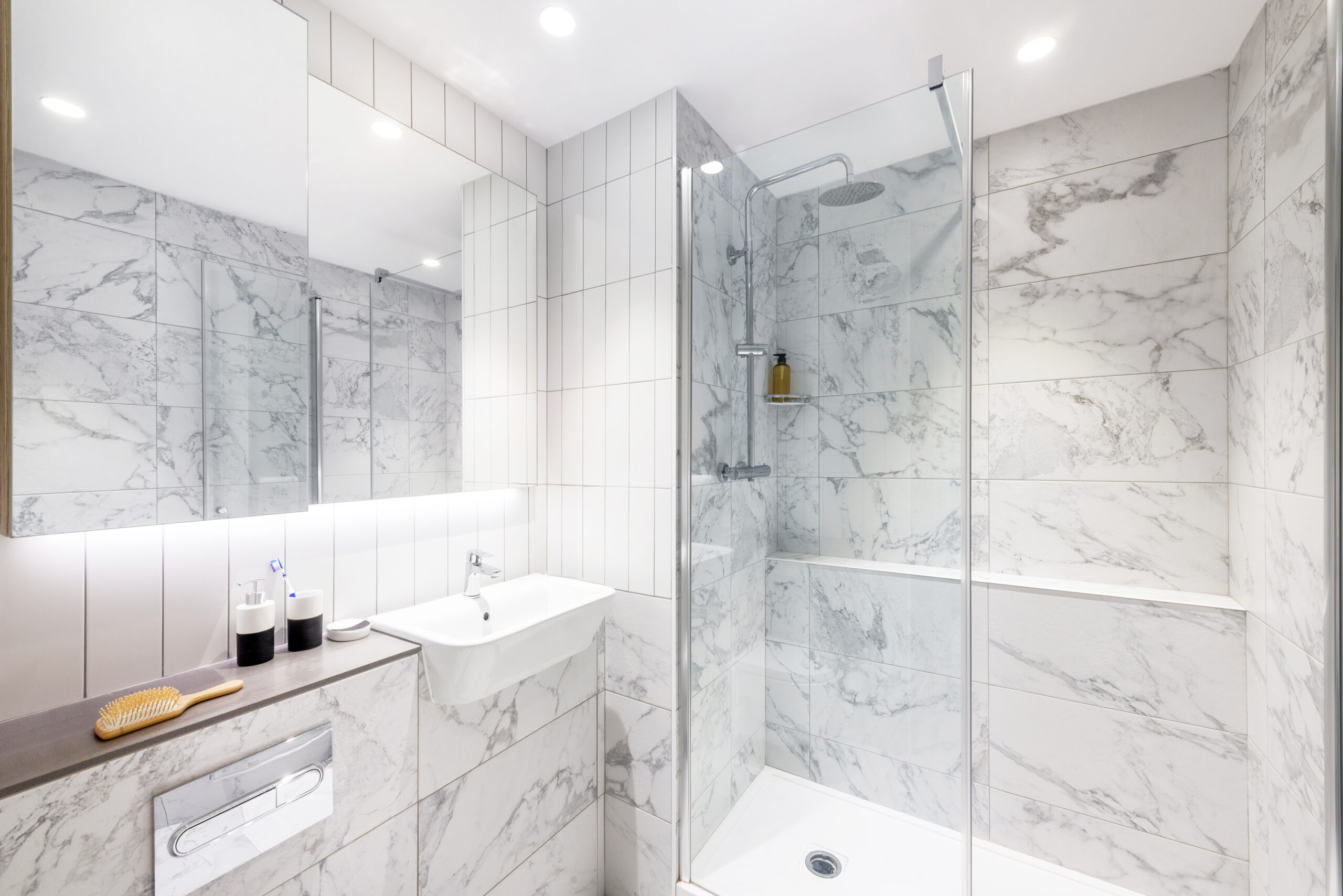 Located on Bruce Street, this modern bathroom boasts marble tiles and a glass-enclosed shower. It features a white sink and mirrored cabinet, while a toothbrush holder and hairbrush sit on the countertop. Recessed lighting brightens the space, enhancing its clean atmosphere.