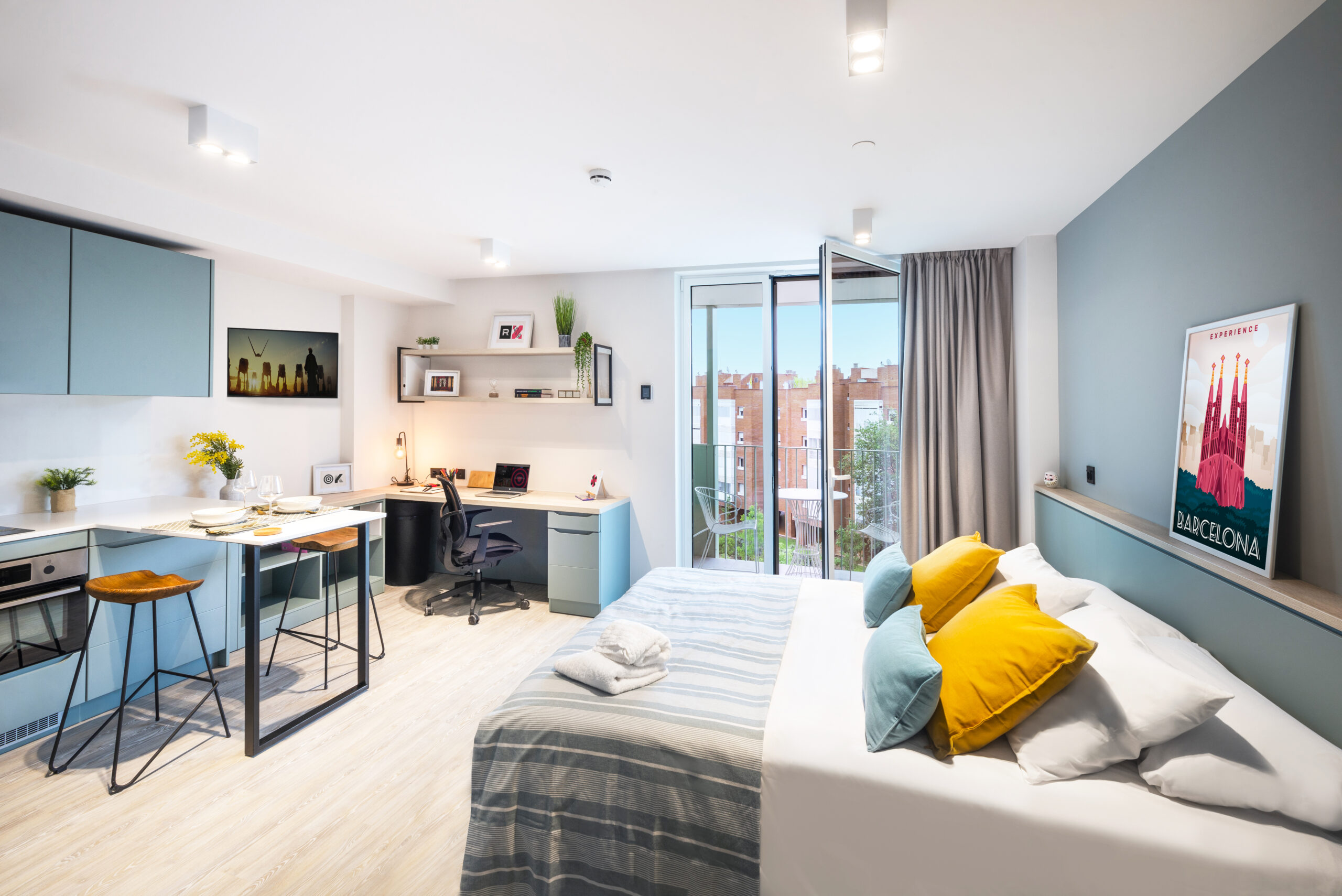 Modern studio apartment on Bruce Street with a cozy bed, colorful pillows, and a striped blanket. The kitchenette features blue cabinets, complemented by a work desk with a chair and a small dining area. A window with sheer curtains offers views of the charming red brick buildings.
