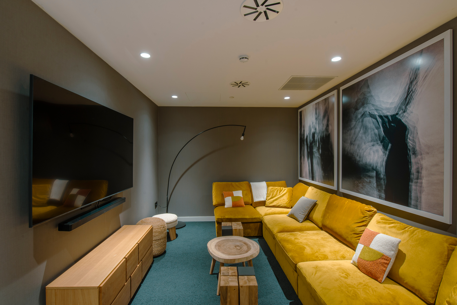 A cozy living room with a mustard-yellow sectional sofa, colorful cushions, and a large flat-screen TV. A round wooden coffee table and modern artwork decorate the space. The room has dim lighting and a minimalist design.