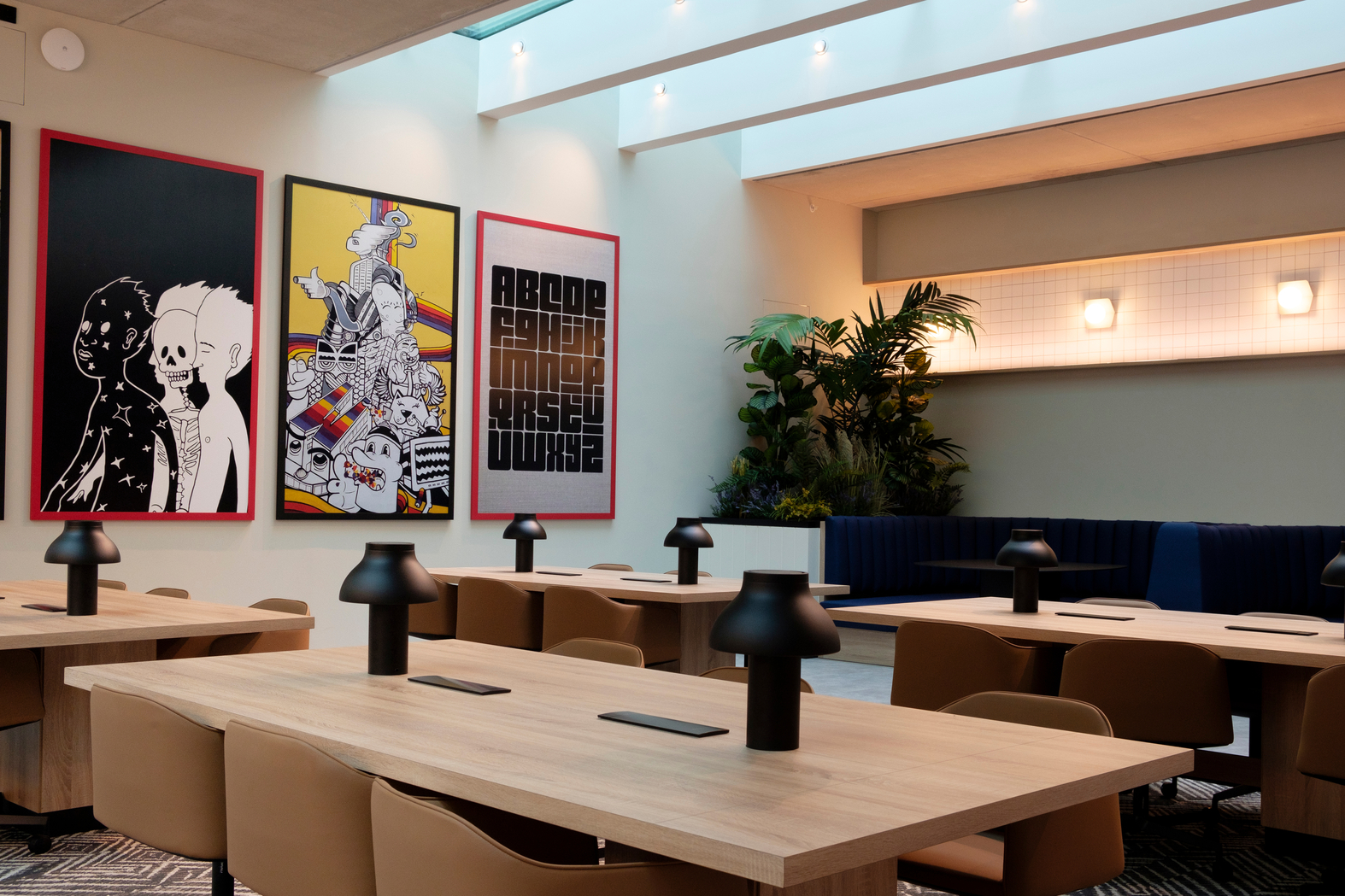 Modern office space with large wooden tables, black desk lamps, and tan chairs. Three colorful art pieces on the wall depict abstract and graphic designs. A skylight above and plants in the corner add natural elements to the room.