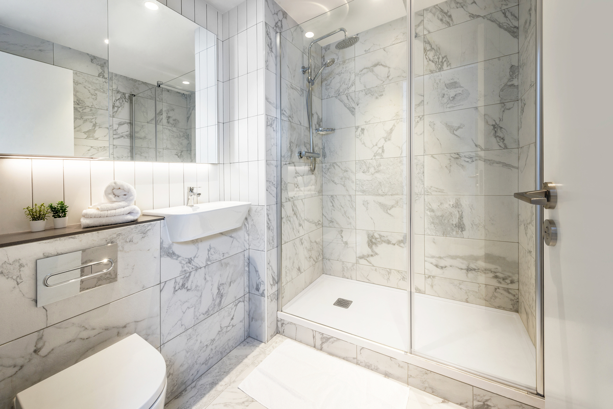 Modern bathroom with marble tiles, a glass-enclosed shower, toilet, wall-mounted sink, large mirror, and countertop with rolled towels and small potted plants. The space is bright and clean with a minimal design.