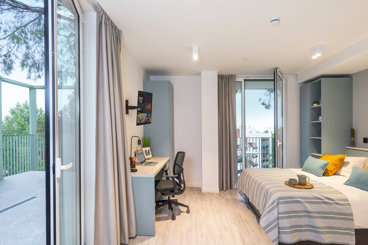 A modern bedroom with a neatly made bed, a desk with a chair and computer, open shelves, and large glass doors leading to a balcony with trees and an outdoor view. The room is bright and spacious.