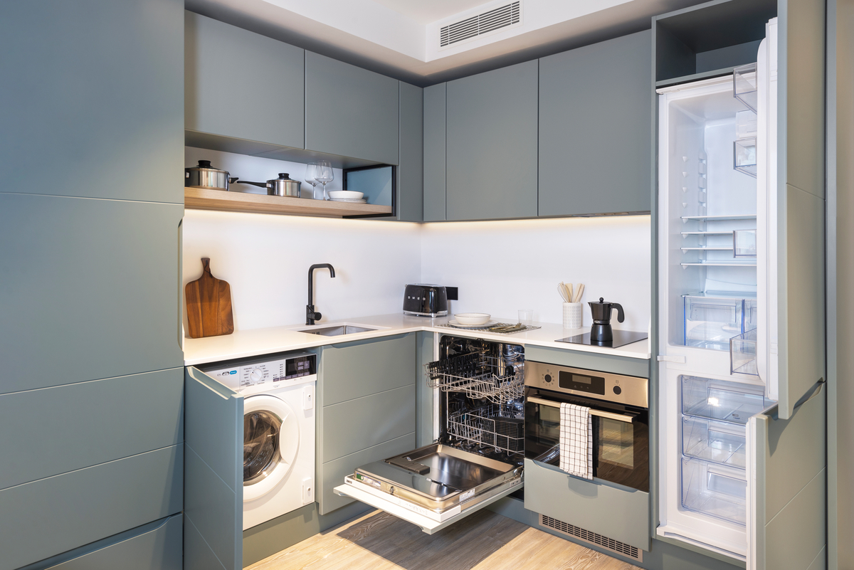 Modern compact kitchen with blue cabinets, open dishwasher, open washing machine, and open refrigerator. Countertop holds a cutting board, utensils, coffee maker, and toaster. Pots and dishes are placed on the upper shelf.