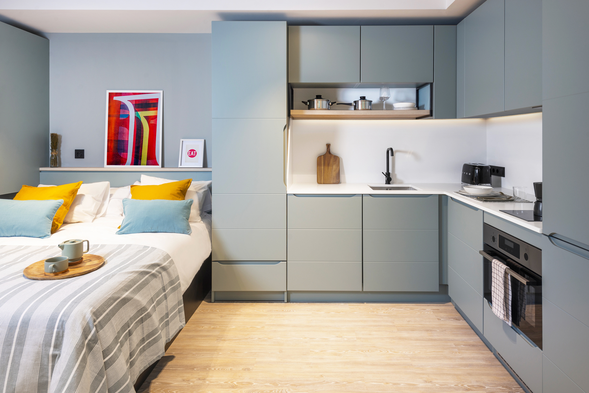 Modern studio apartment with a sleek grey kitchen, built-in appliances, and a bed with pillows and a striped blanket. Colorful artwork and a breakfast tray add accents; light wooden floor brightens the space.