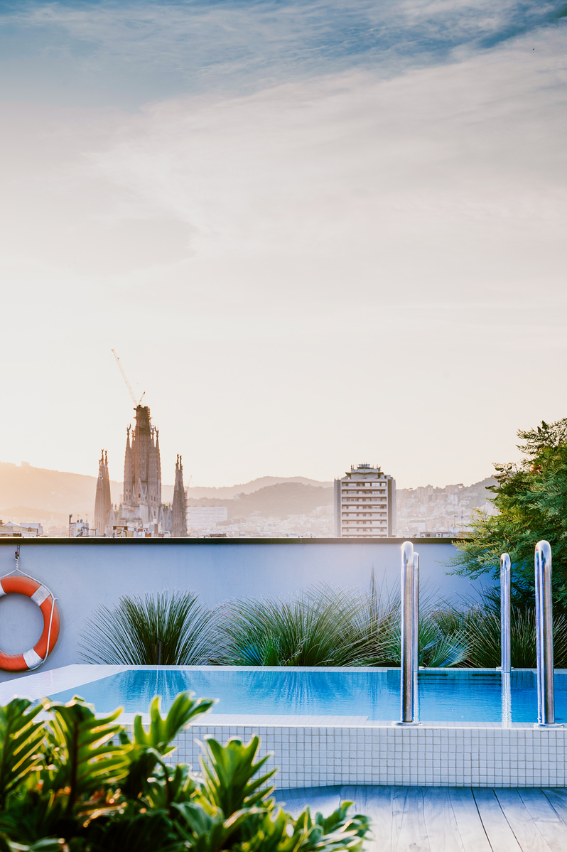A rooftop pool with metal handrails overlooks the city of Barcelona, featuring the Sagrada Família basilica and surrounding buildings under a soft, cloudy sky at sunset.