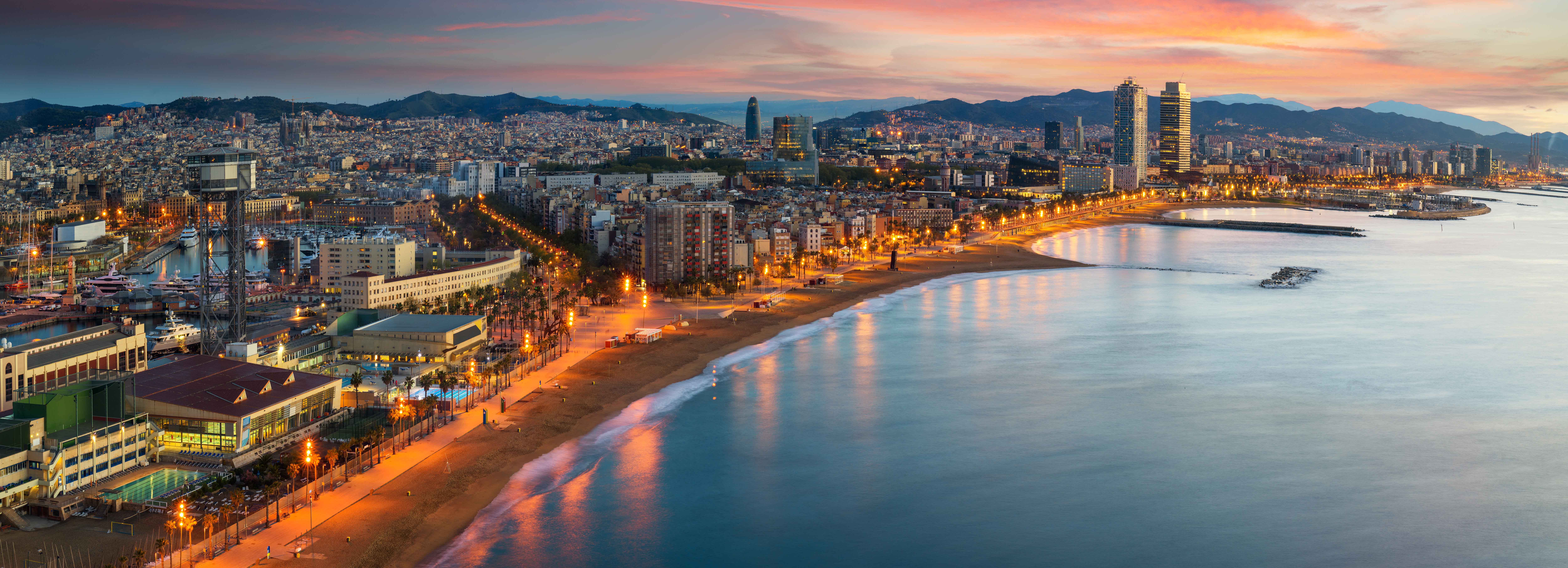 Panoramic view of a coastal city at sunset, featuring a wide sandy beach with calm waters. The skyline is dotted with buildings, and the scene glows with city lights. Mountains are visible in the background under a colorful sky.