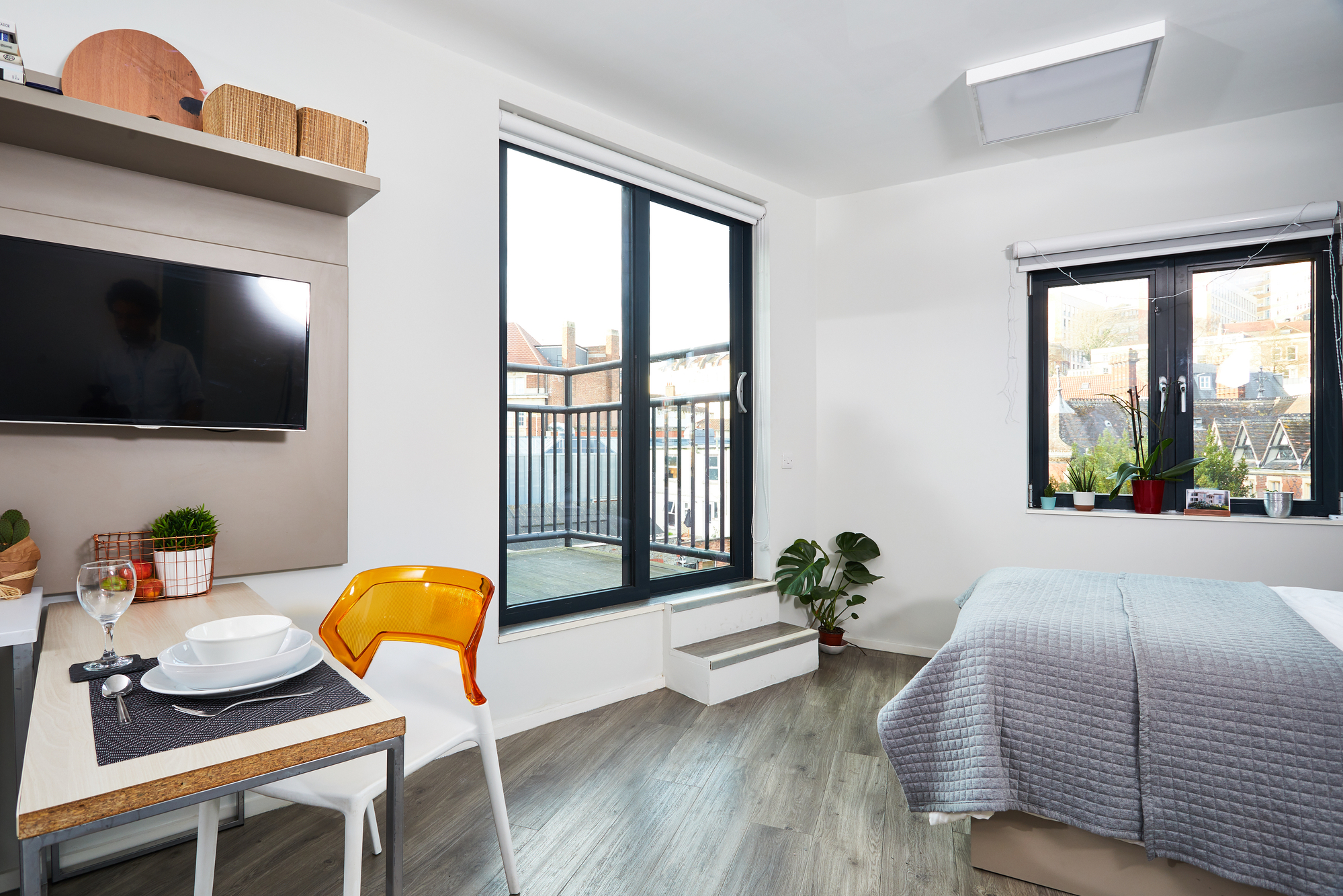 A modern studio apartment with a bed, a small dining table set for two, a wall-mounted TV, large windows, and a sliding door leading to a balcony. The room features plants and contemporary decor.