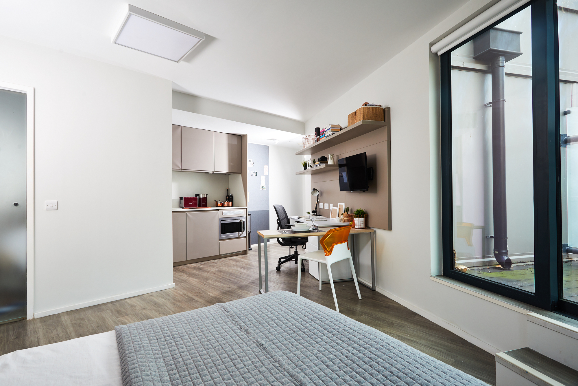 A modern studio apartment with a bed, a desk and chair, shelves, a mounted TV, and a compact kitchen. Large windows let in natural light, and the decor is minimalist with light-colored walls and wood flooring.
