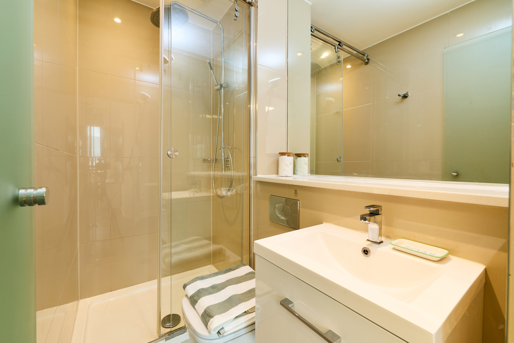 Modern bathroom with a glass shower enclosure, striped towel on the shower floor, wall-mounted toilet, white vanity with sink and soap, and a large mirror reflecting the room. Neutral beige and white color palette.