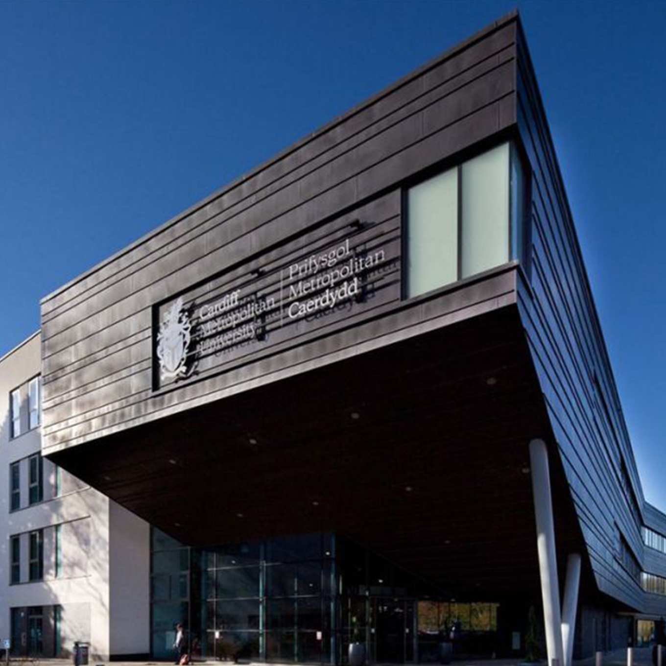 Modern building with large overhang and dark facade, displaying the Cardiff Metropolitan University logo and Welsh text. Clear blue sky in the background.