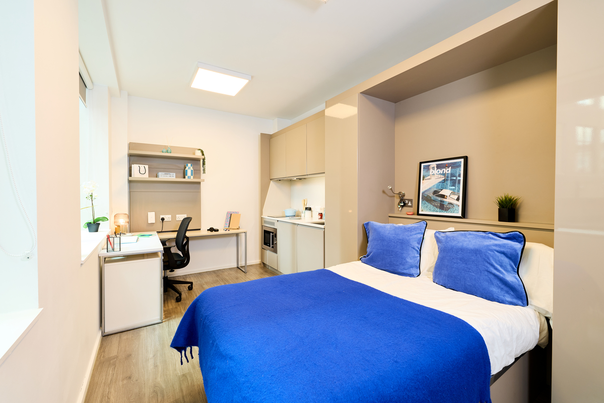 Bright, modern student bedroom with a double bed covered in blue bedding, a desk with a chair and lamp near the window, shelves, a small kitchenette, and wall art above the bed.