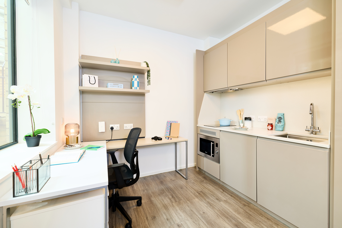 A modern studio apartment with a compact kitchen, a desk and chair by the window, open shelves with books and decorations, and wood-style flooring. The space is bright and neatly organized.