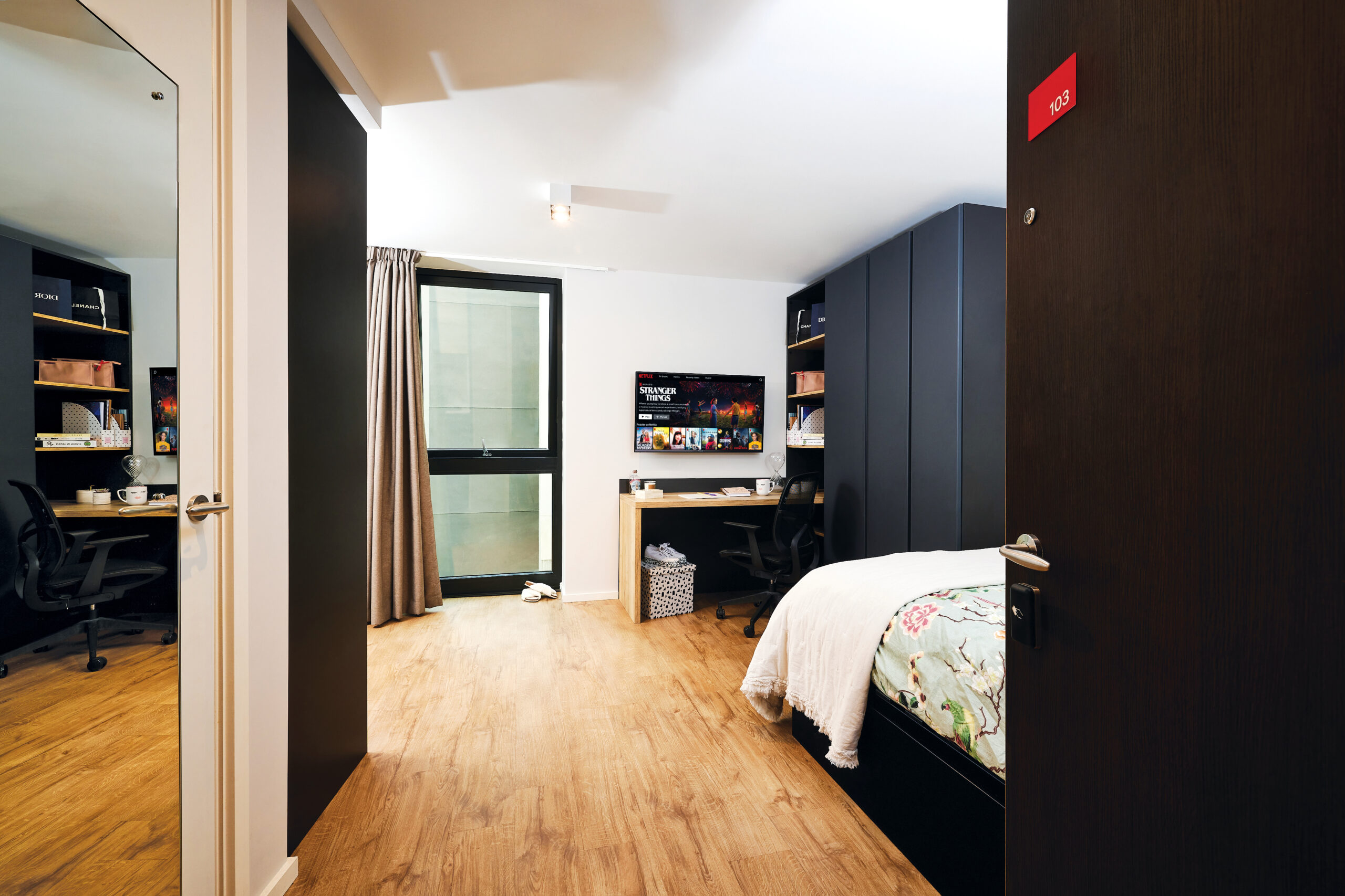 Modern dorm room with wooden flooring in Portland, featuring a bed with a colorful blanket on the right, a desk with a computer against the far wall, and a window with beige curtains. A large mirror and a black door with a crescent-shaped red room number are visible.