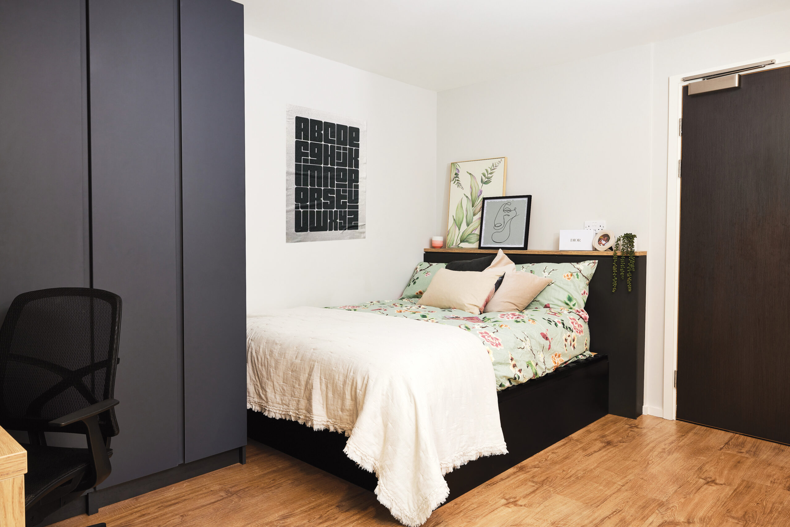 Cozy bedroom on Portland Crescent featuring a neatly made bed with floral bedding and a white throw blanket. A small plant and framed artwork adorn the headboard shelf. On the left, a dark wardrobe and office chair sit, while a closed door is on the right.