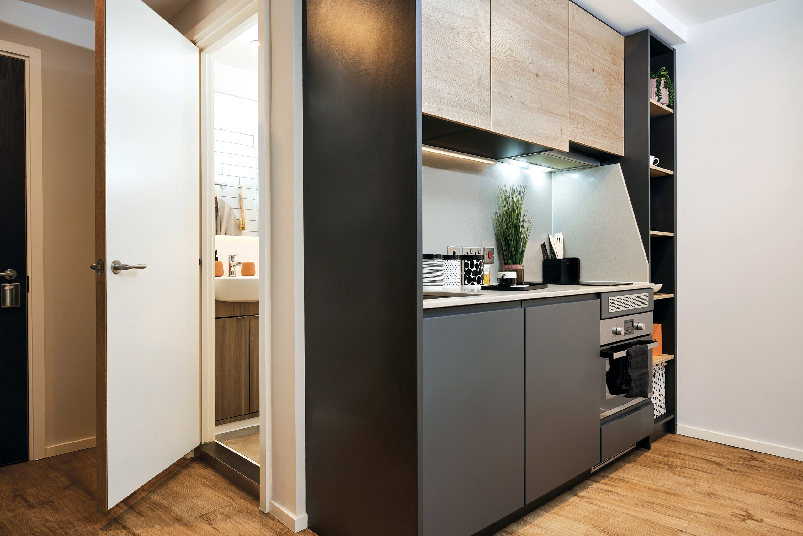 Compact modern kitchen in Portland with wood and dark cabinetry, featuring a Crescent sink, countertop, oven, and overhead storage. Open door reveals a small bathroom with a sink and mirror. Shelving contains decorative items. Light wooden flooring throughout.