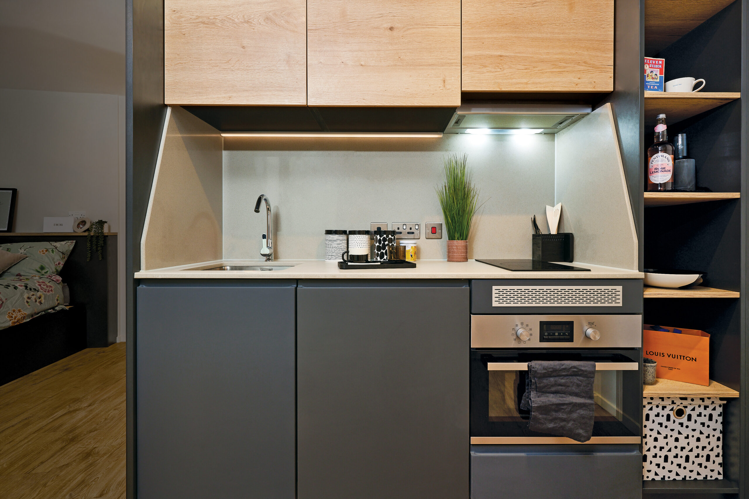 A modern kitchenette in Portland features wood and grey cabinetry, a sink, oven, and induction cooktop. Accessories like a potted plant, spice jars, and a towel on the oven handle add charm. Crescent shelving on the right holds a cup and other items with the bedroom partially visible.