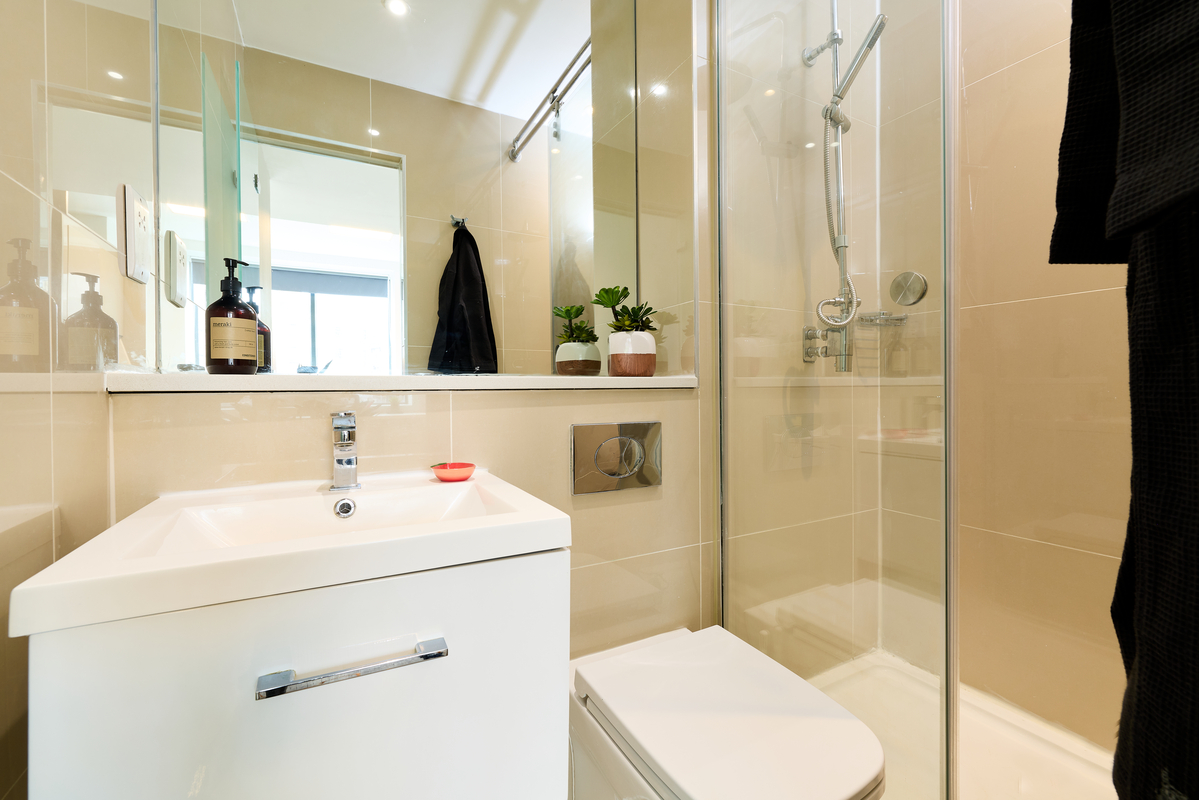Modern bathroom with a white sink, mirror, toilet, glass-enclosed shower, beige tiled walls, and small potted plants on a shelf. Soap dispenser and towel are visible, with natural light coming in.