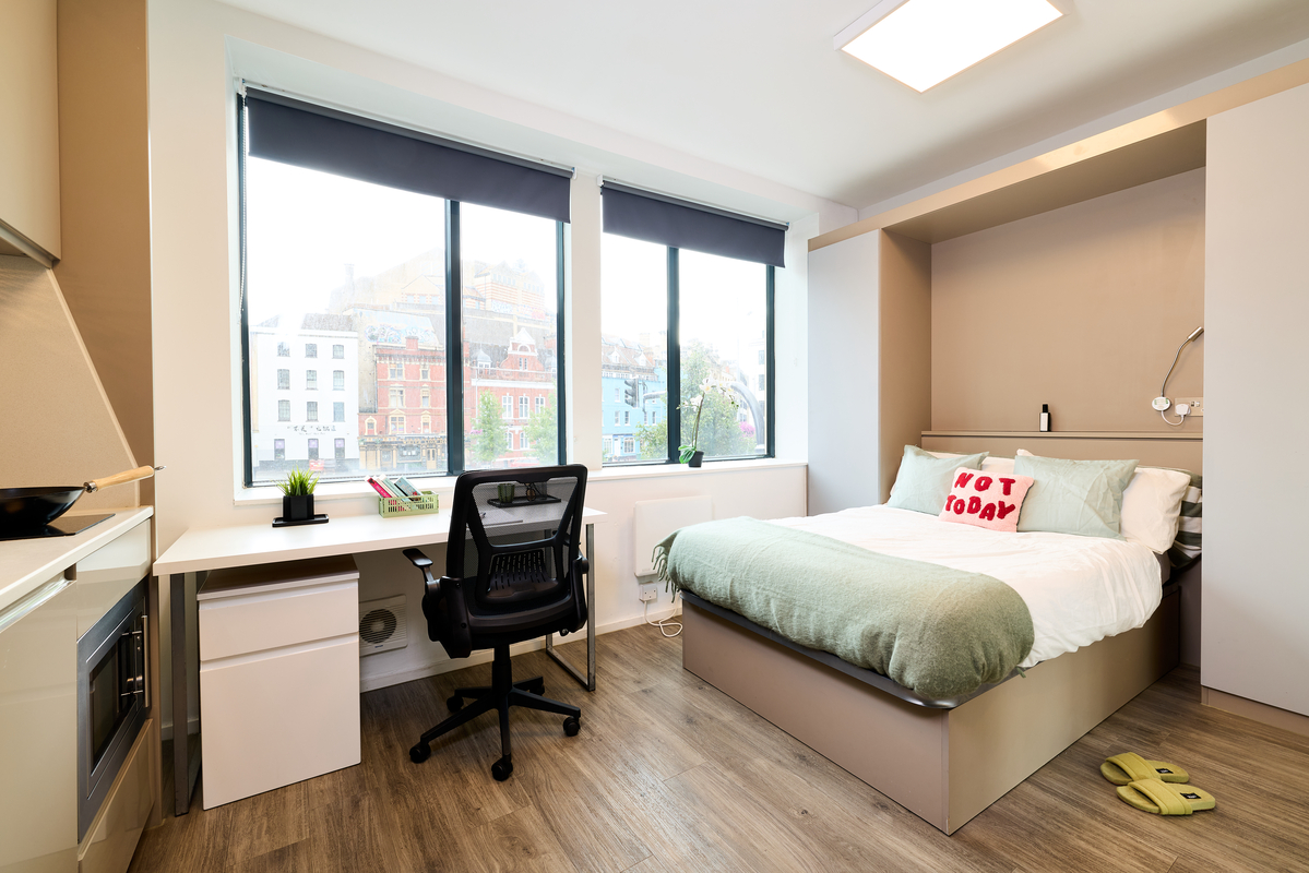 A modern, bright dorm room with large windows, a desk and chair, a neatly made bed with a "Not Today" pillow, slippers on the floor, and a small kitchenette visible on the left.