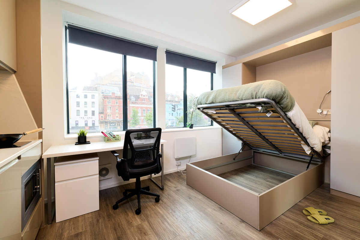 A modern studio room with wood floors features a large window, a desk with a black chair, and a raised storage bed with sheets and slippers beside it. Buildings are visible outside the window.