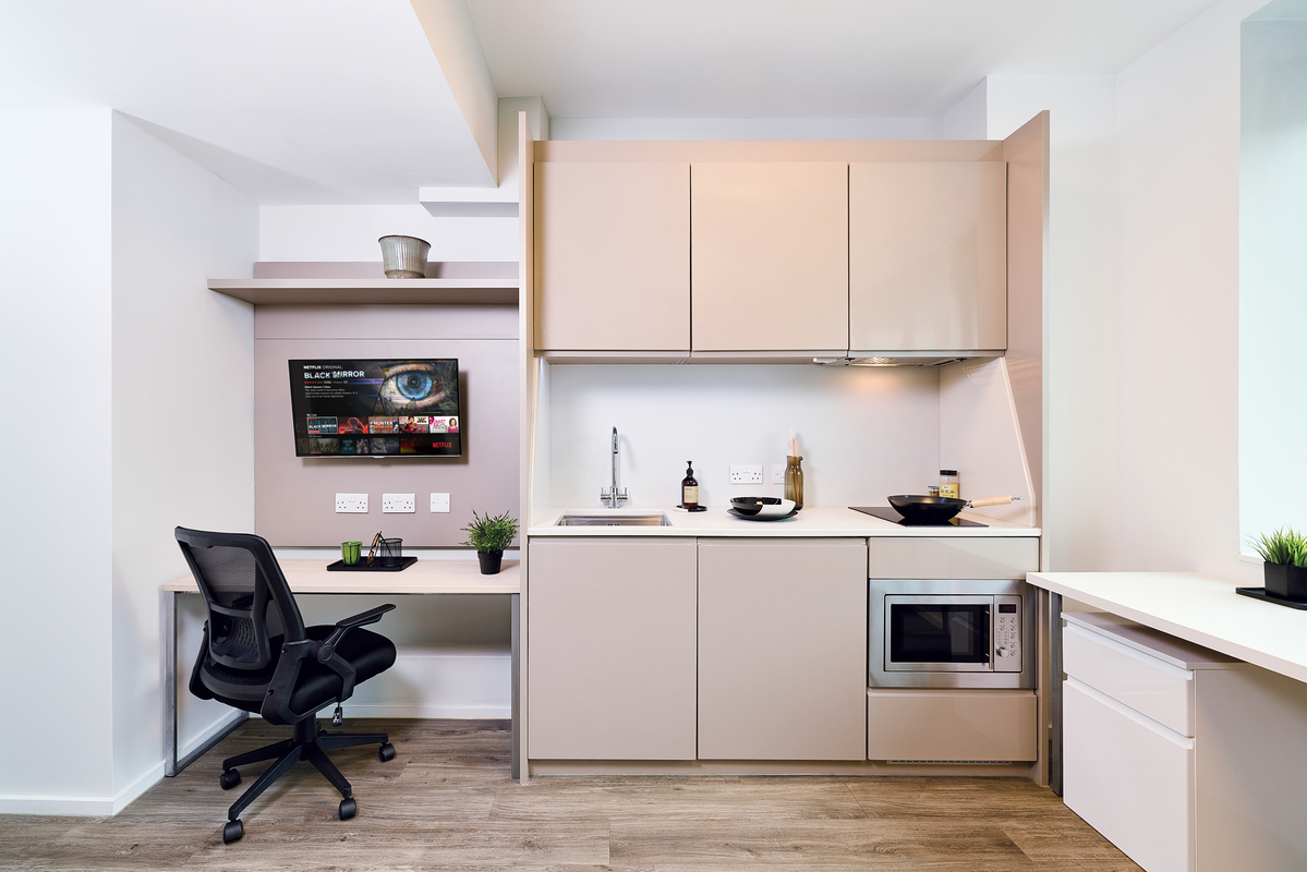 Modern studio apartment with a compact kitchen, built-in microwave, sink, stovetop, and beige cabinets. A work desk with a black chair and wall-mounted TV is on the left. Green plants add a touch of decor.