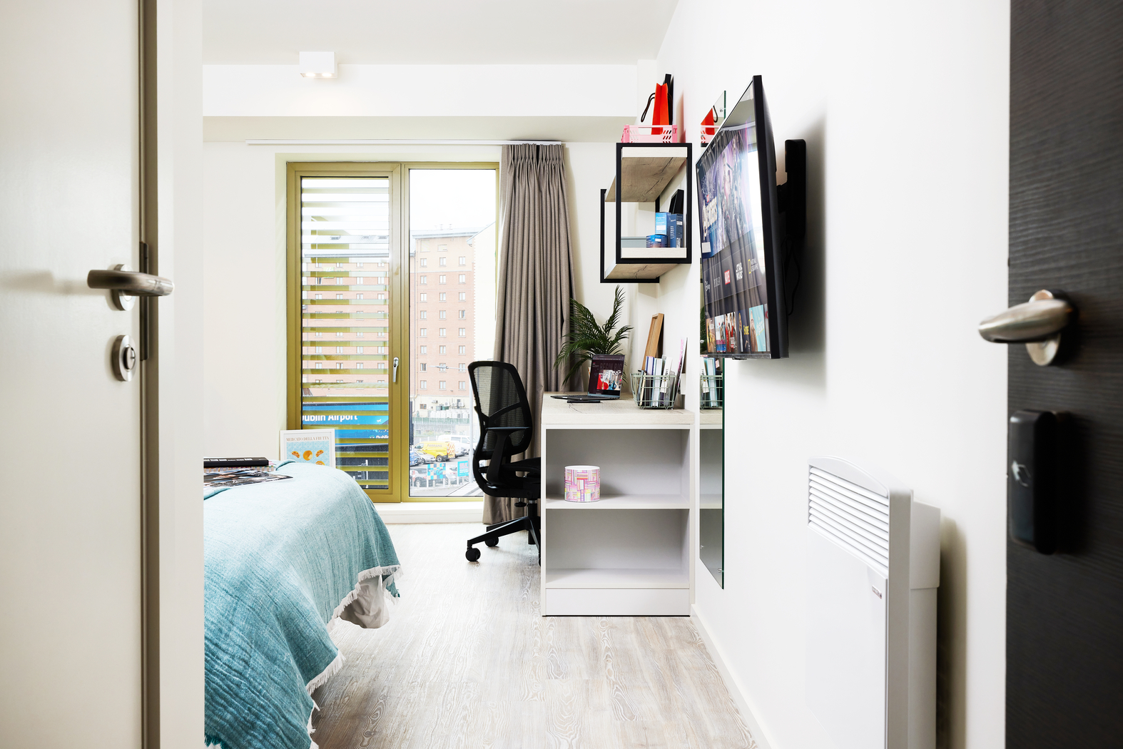 Nestled on Bruce Street, this modern bedroom features a large bed draped in a blue blanket. A desk with a black chair faces the wall-mounted TV, while shelves hold books and decor. A window with blinds and curtains invites natural light to enhance the room's ambiance.