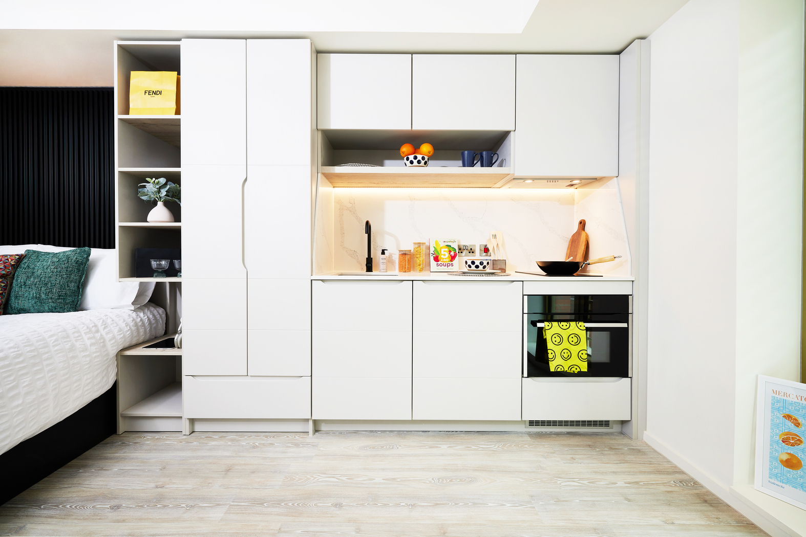 Modern studio apartment kitchen on Bruce Street with white cabinets, built-in oven, and minimalist decor. A small plant, bowls, and kitchen essentials are neatly arranged. Light wood flooring complements the bright, clean aesthetic. A bed is partially visible on the left.