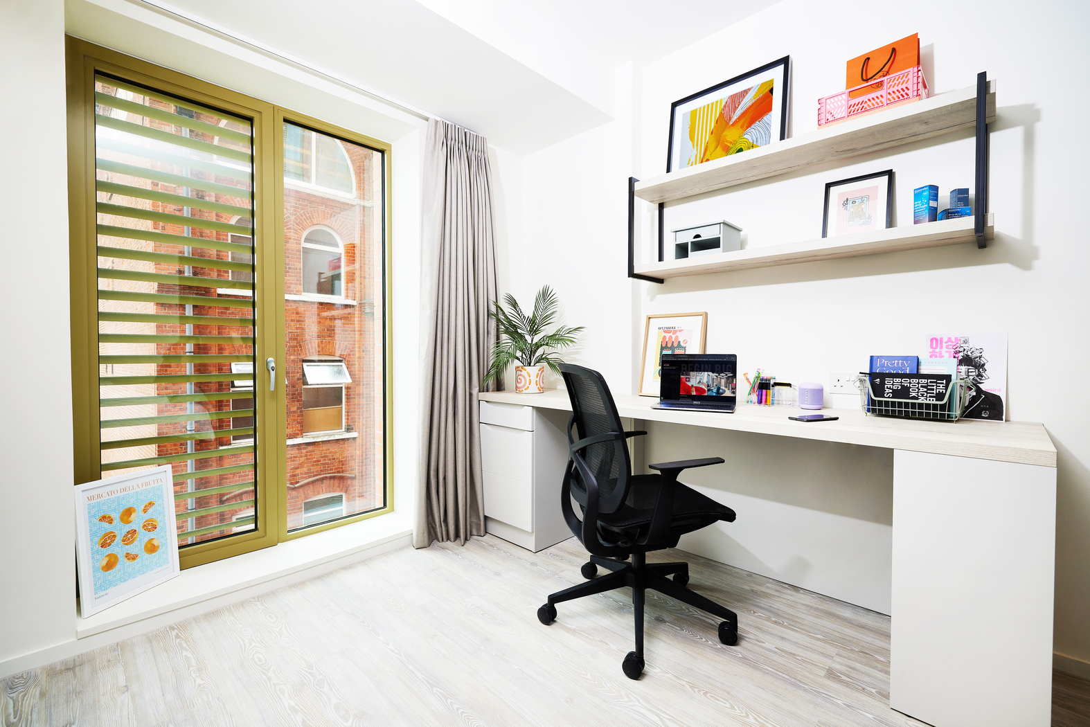 Located on Bruce Street, this bright home office features a large window, desk, and office chair. You'll find a laptop, headphones, framed art, and stationery on the desk. A plant rests on a side table while wall shelves showcase books and decorative items. Light wooden flooring enhances the space.