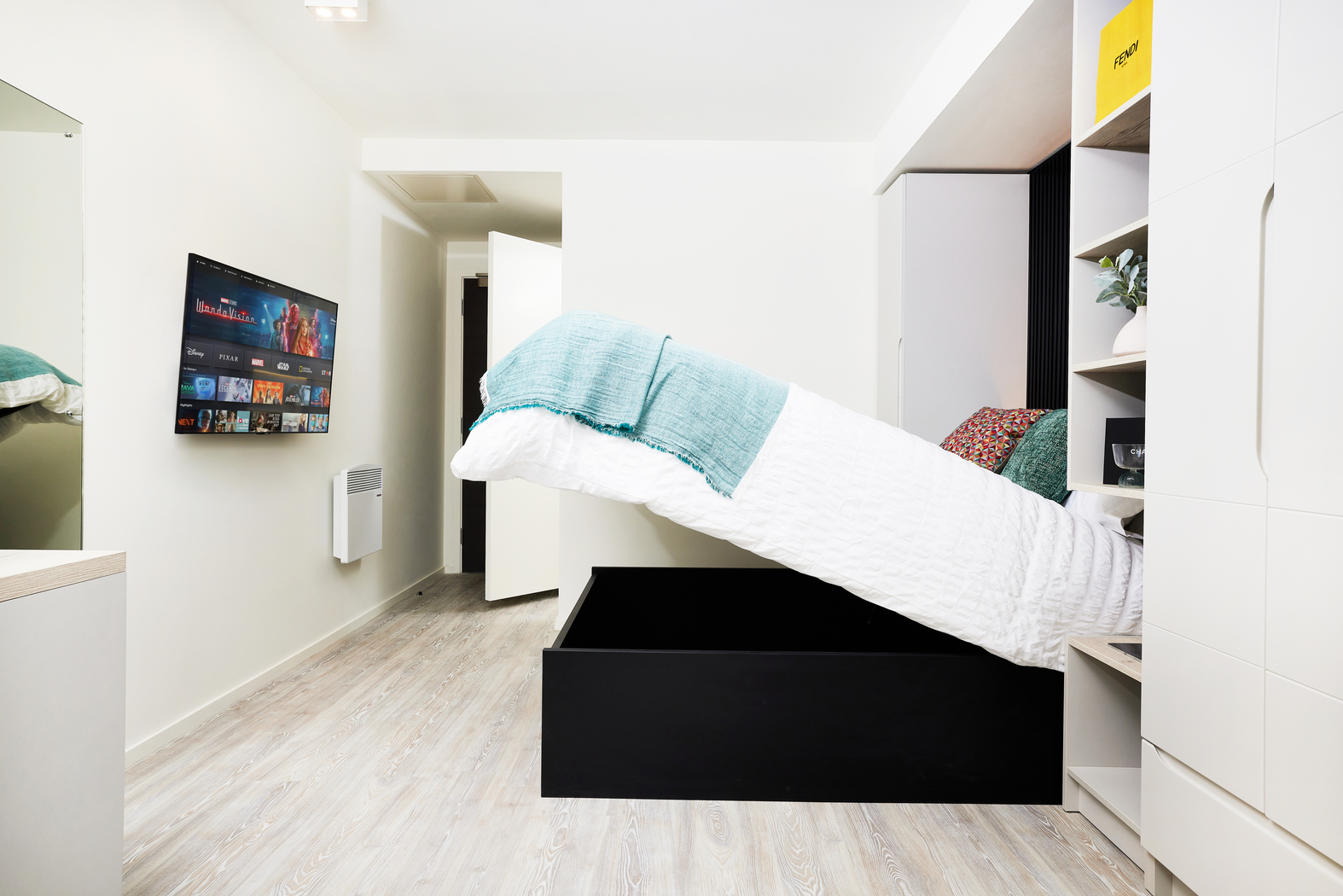 On Bruce Street, a minimalist bedroom features a lifted storage bed with white bedding and teal pillows, revealing space underneath. A wall-mounted TV graces the left wall, while light wood flooring complements the white walls. A tall wardrobe stands on the right.