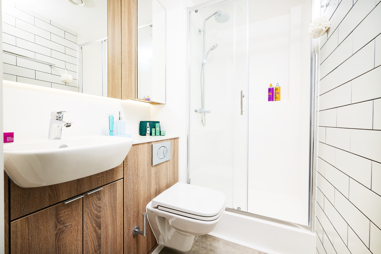 Located on Bruce Street, this modern bathroom boasts white tiles and wood accents. It features a sleek glass shower, a white sink with storage beneath, and a floating toilet. Toiletries are neatly arranged on the counter and in the shower for convenience.