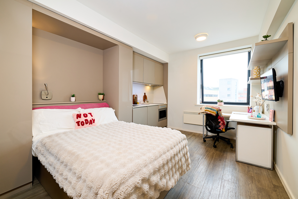 Modern, tidy student bedroom in Portland House with a double bed, "Not Today" pillow, kitchenette, desk with computer, shelving, large window, and light wooden floor. The décor is minimalist with neutral tones and small plants.
