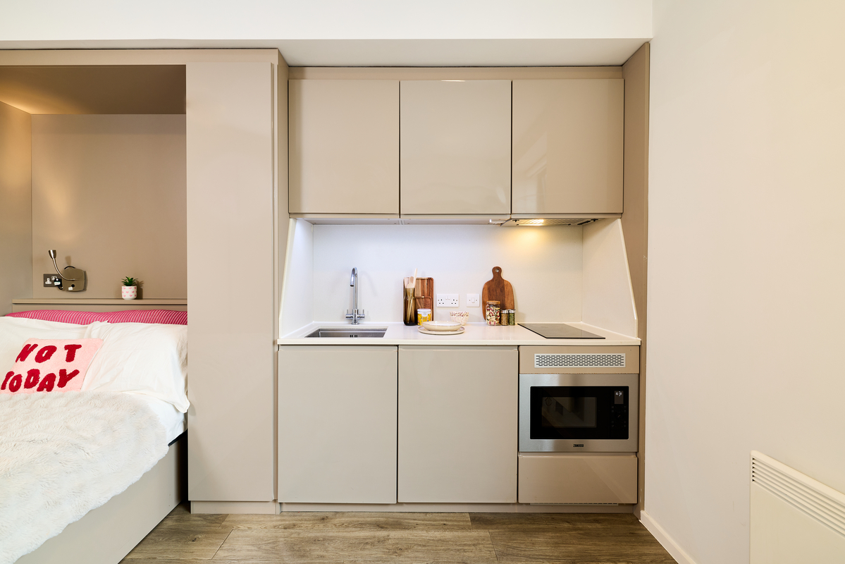 Modern compact kitchen at Portland House with beige cabinets, a sink, electric stovetop, microwave, and wooden floor. A neatly made bed featuring a "NOT TODAY" pillow is partially visible on the left side.