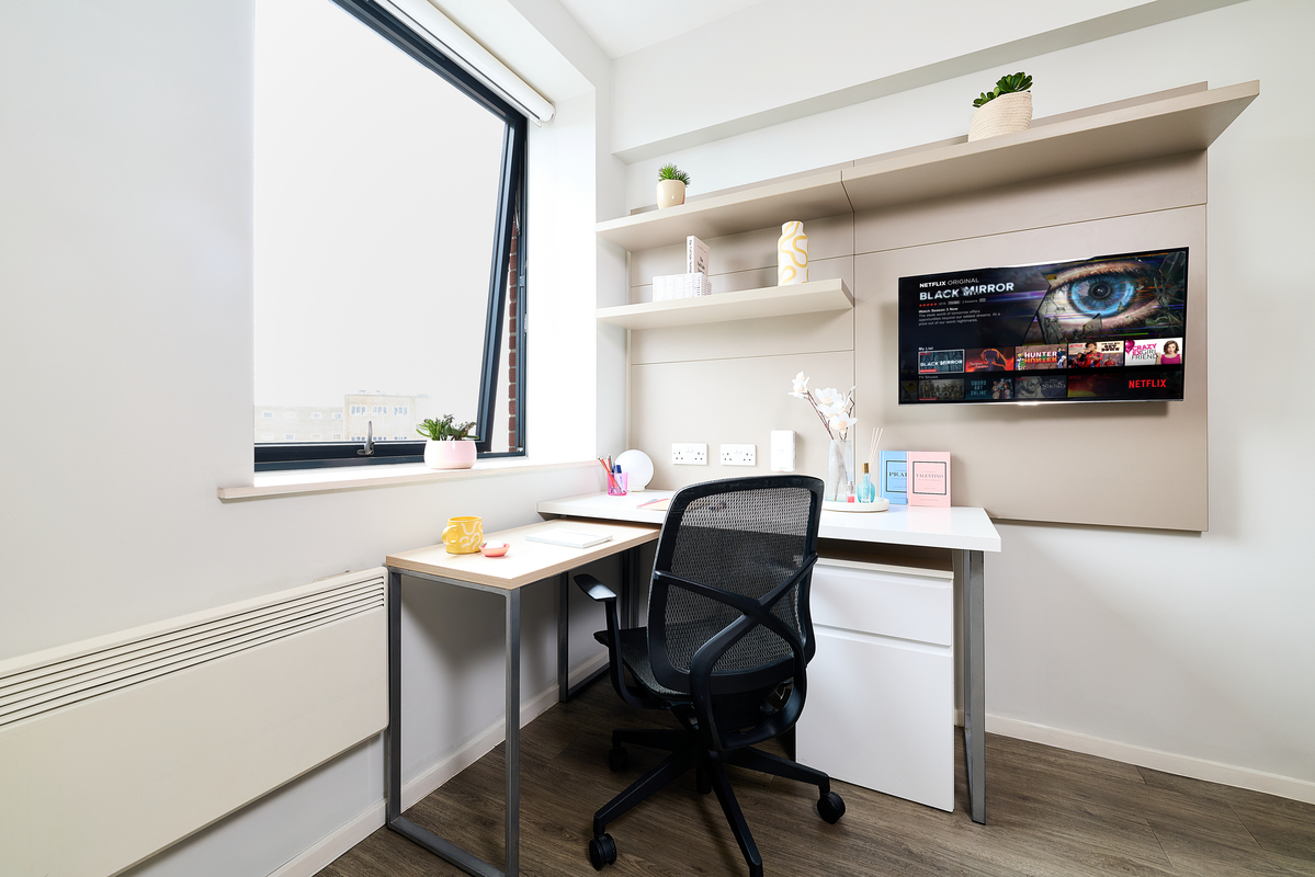 Modern, tidy workspace at Portland House with a desk, black chair, and shelving. A TV displaying Netflix is mounted on the beige wall. Large window, indoor plants, and soft lighting create a bright, inviting atmosphere.