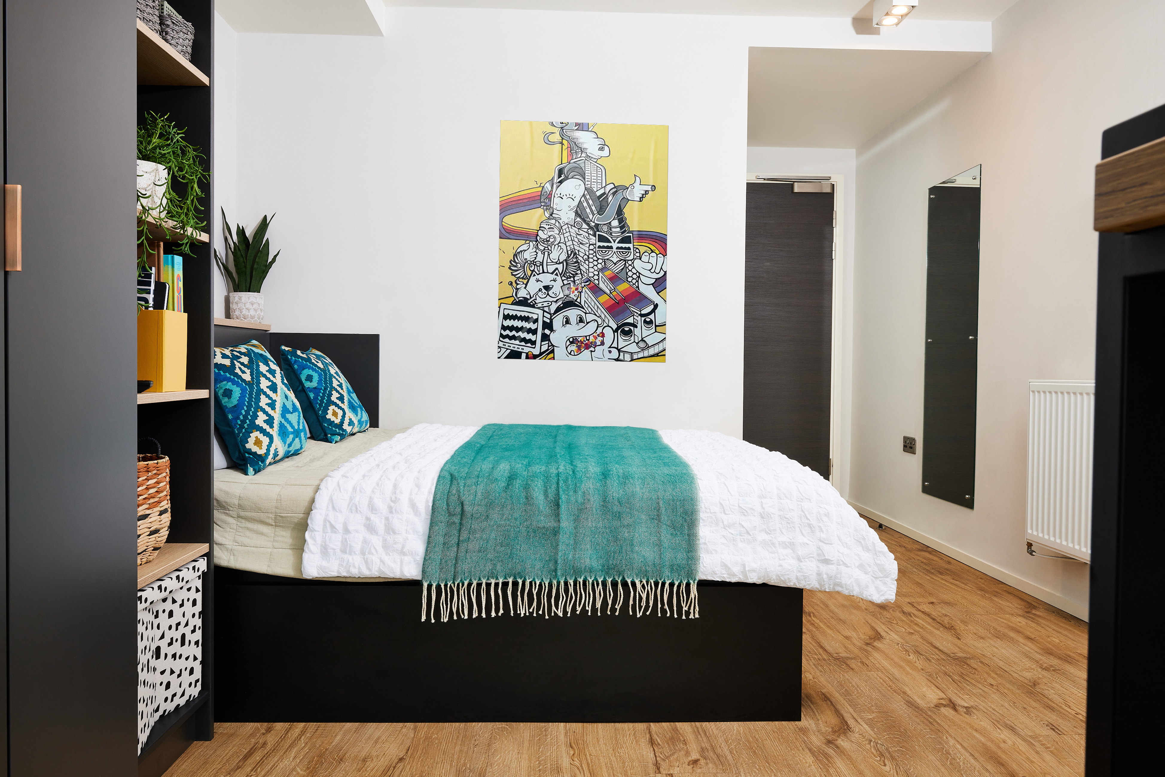 A modern bedroom with a neatly made bed featuring turquoise pillows and a throw. The wall has a colorful abstract painting. Shelves with plants and baskets are on the left. The room has wooden flooring and a full-length mirror on the right.