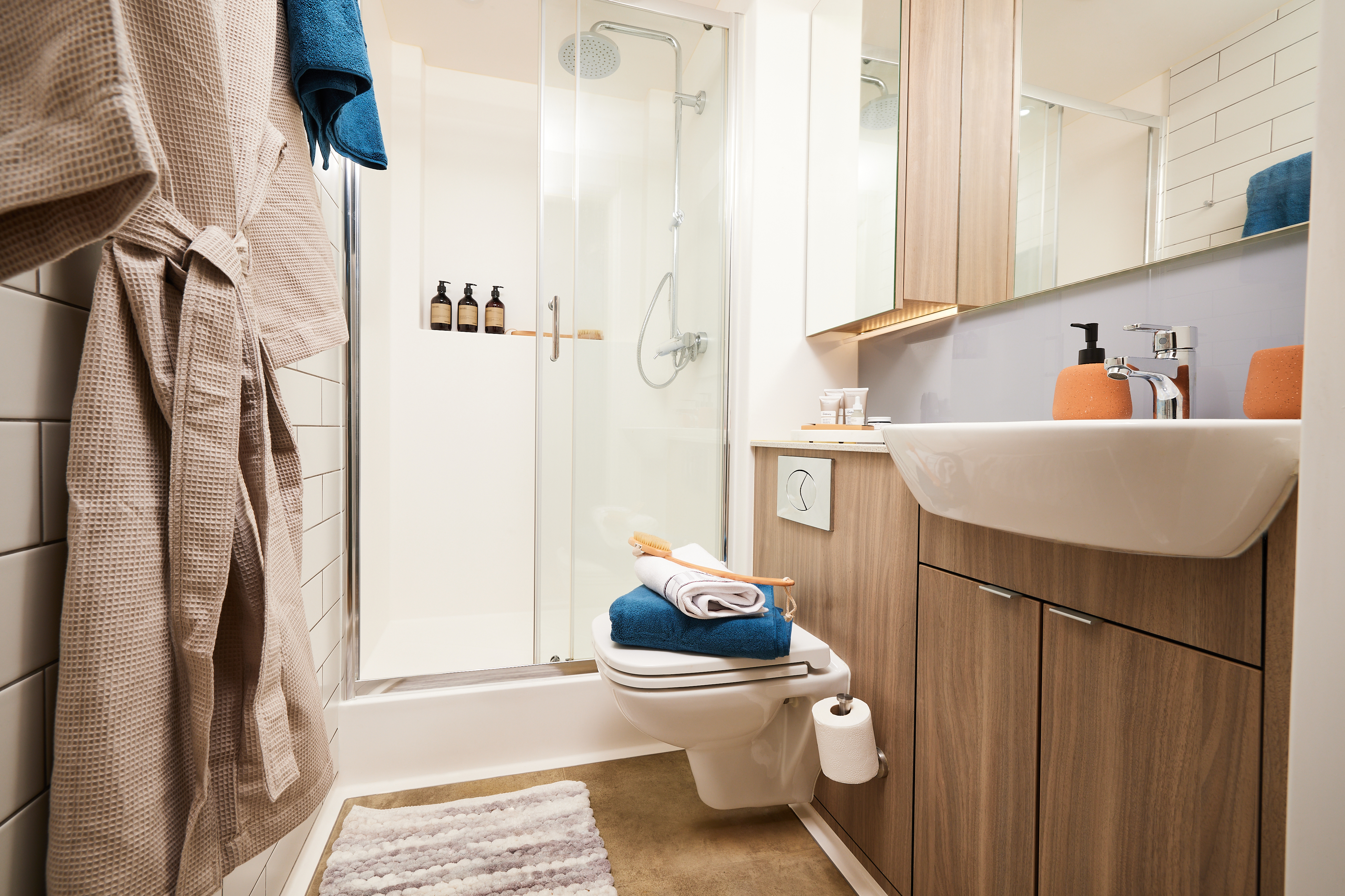 Modern bathroom with a walk-in shower, glass doors, and wooden cabinetry. A sink is mounted on the right with toiletries on the counter. Towels and a washcloth rest on the toilet. A robe hangs on the left wall, and a striped rug lies on the floor.