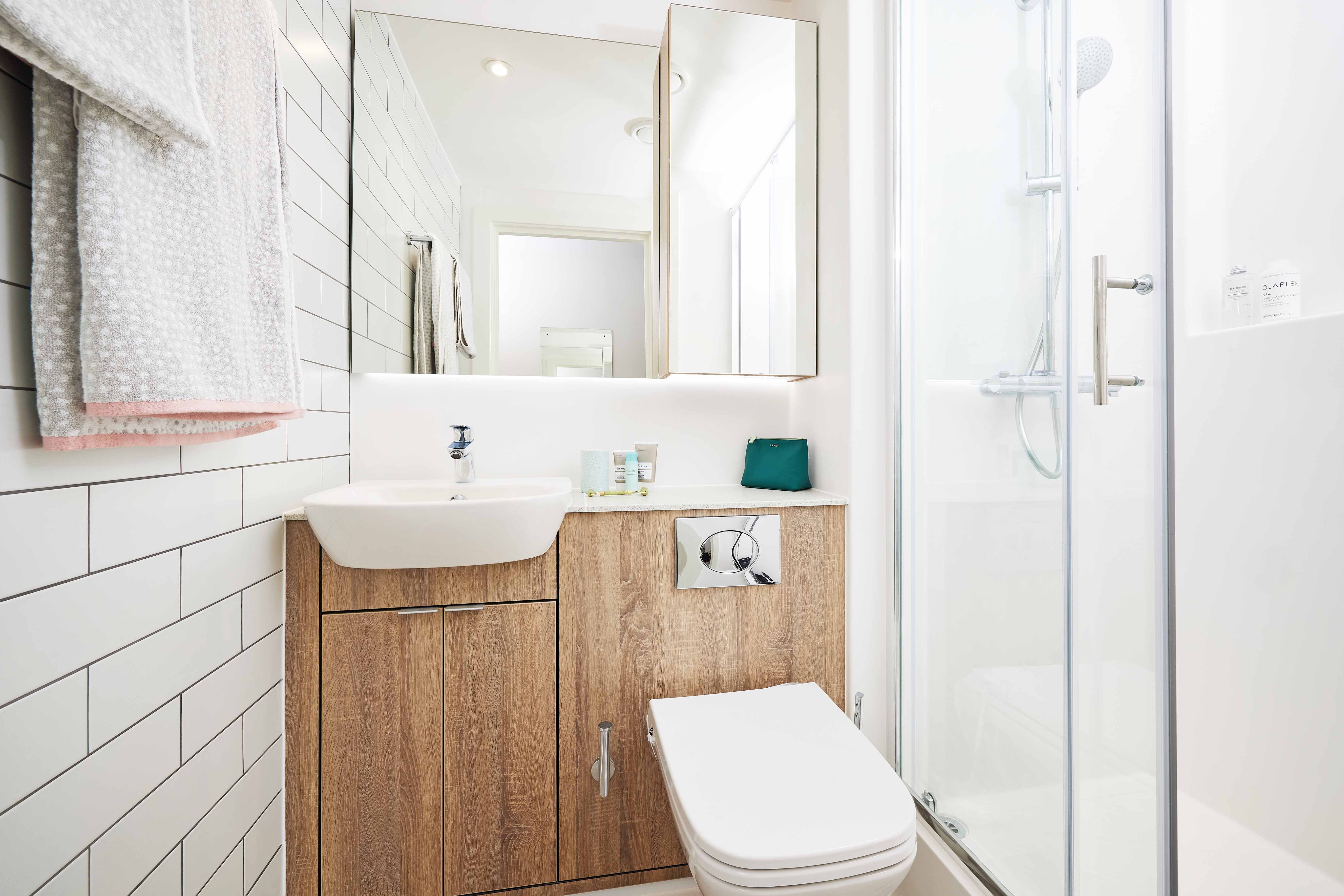 Modern bathroom with a wooden cabinet, white sink, and wall-mounted faucet. A glass shower enclosure is on the right, and white tiled walls add brightness. A mirror and folded towels are visible, with a small window providing natural light.
