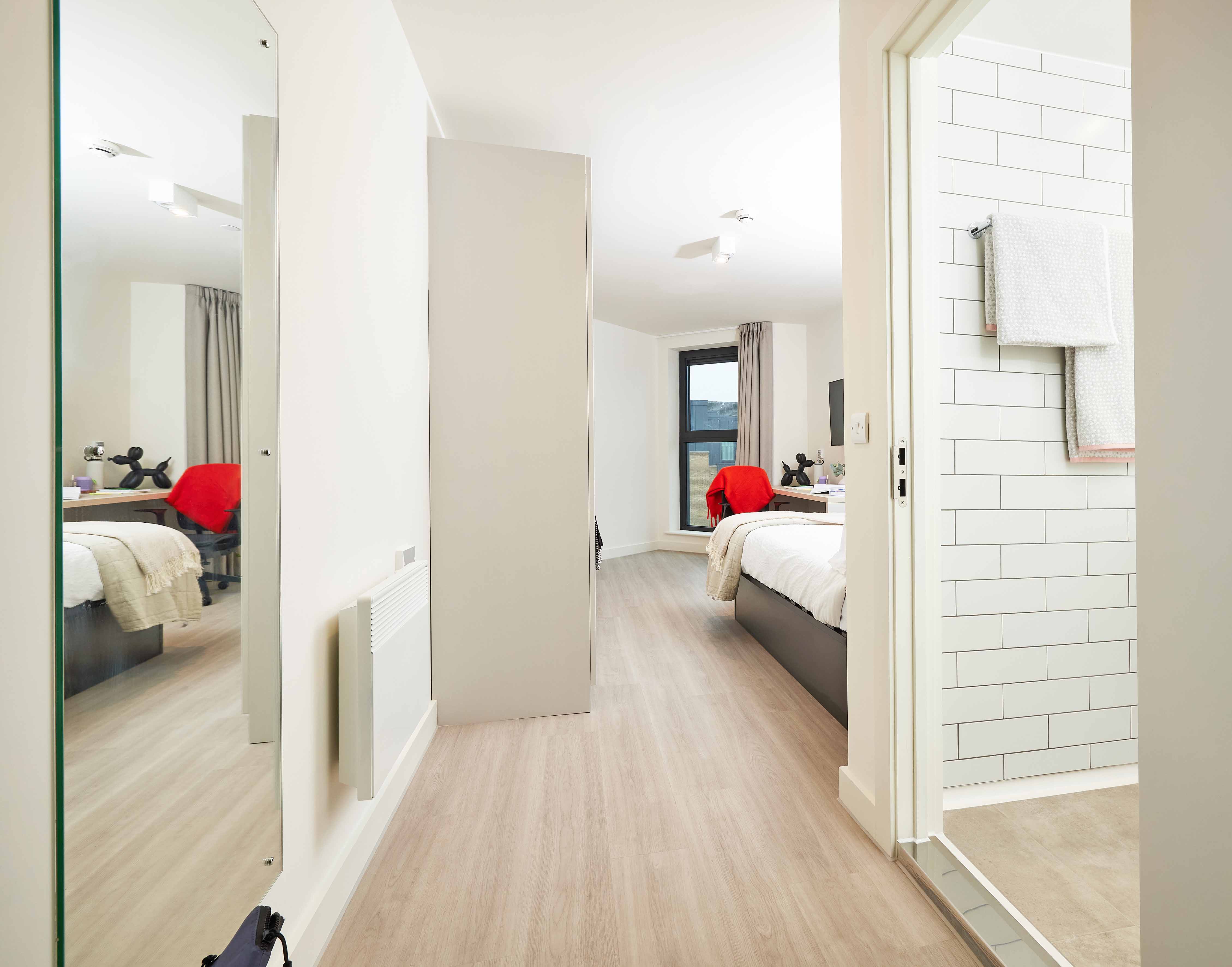 View of a modern, clean bedroom with a neatly made bed and a red throw. A large mirror reflects the room, and an adjacent bathroom features white tile walls. Light wood flooring runs throughout the space.