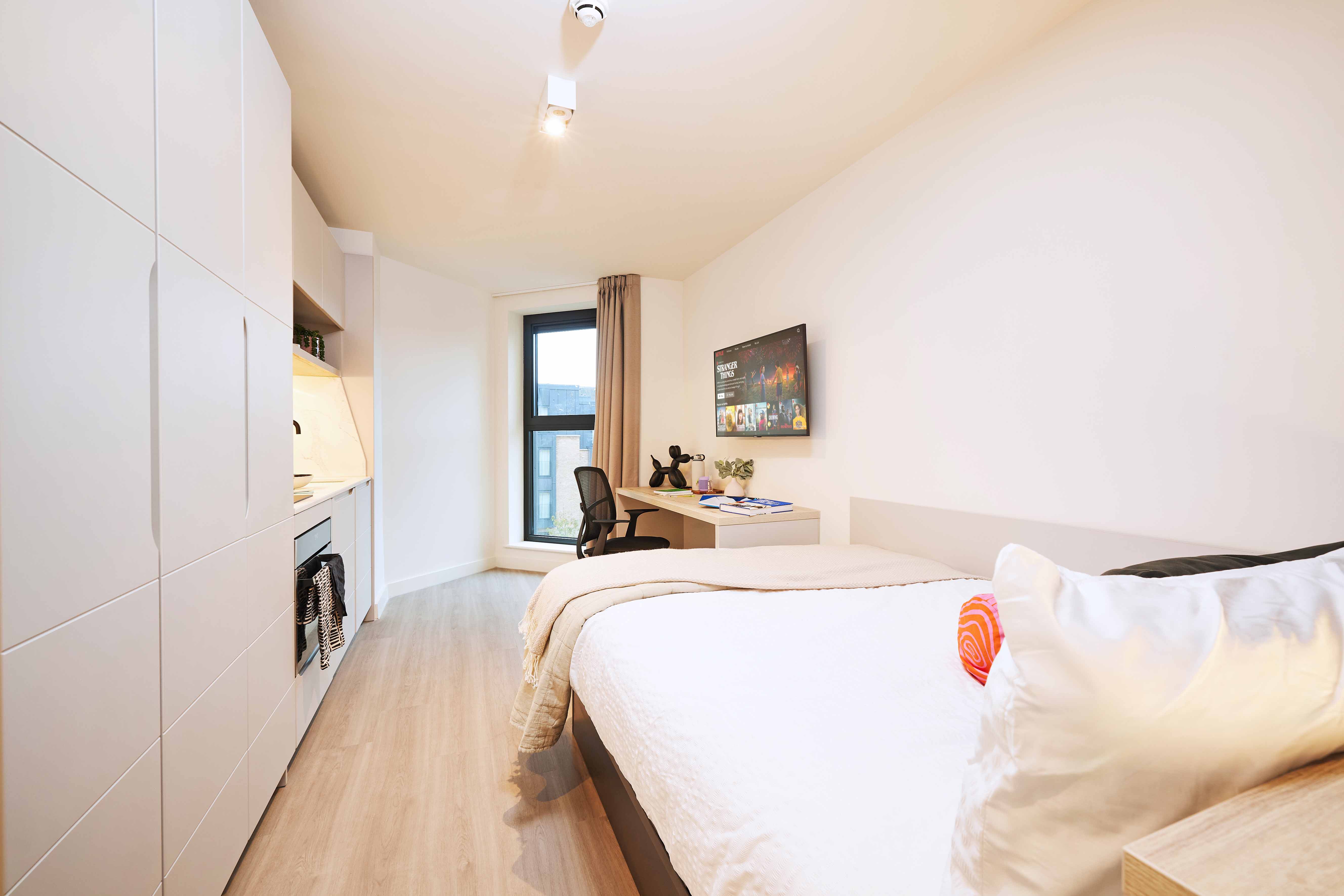 Modern, compact bedroom with a neatly made bed facing a wall-mounted TV. A kitchenette with cabinets is on the left, and a window with blinds lets in natural light. A desk beside the bed holds books and decor. The space is bright and minimalistic.