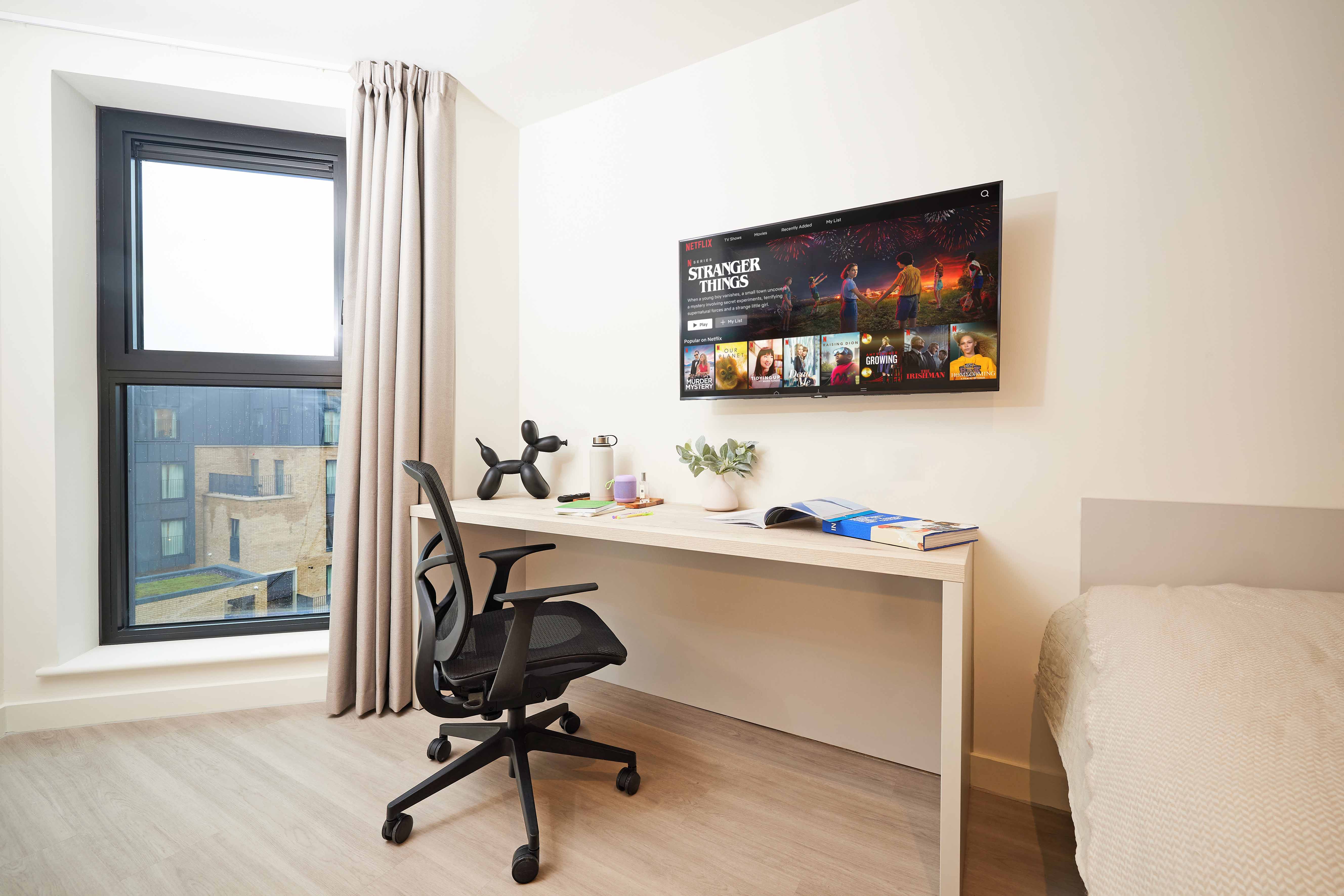 Modern bedroom with a desk and ergonomic chair, a TV mounted on the wall displaying a streaming service, a bed with white linens, a large window with closed curtains, and various desk items including books, a houseplant, and a unique dog-shaped sculpture.