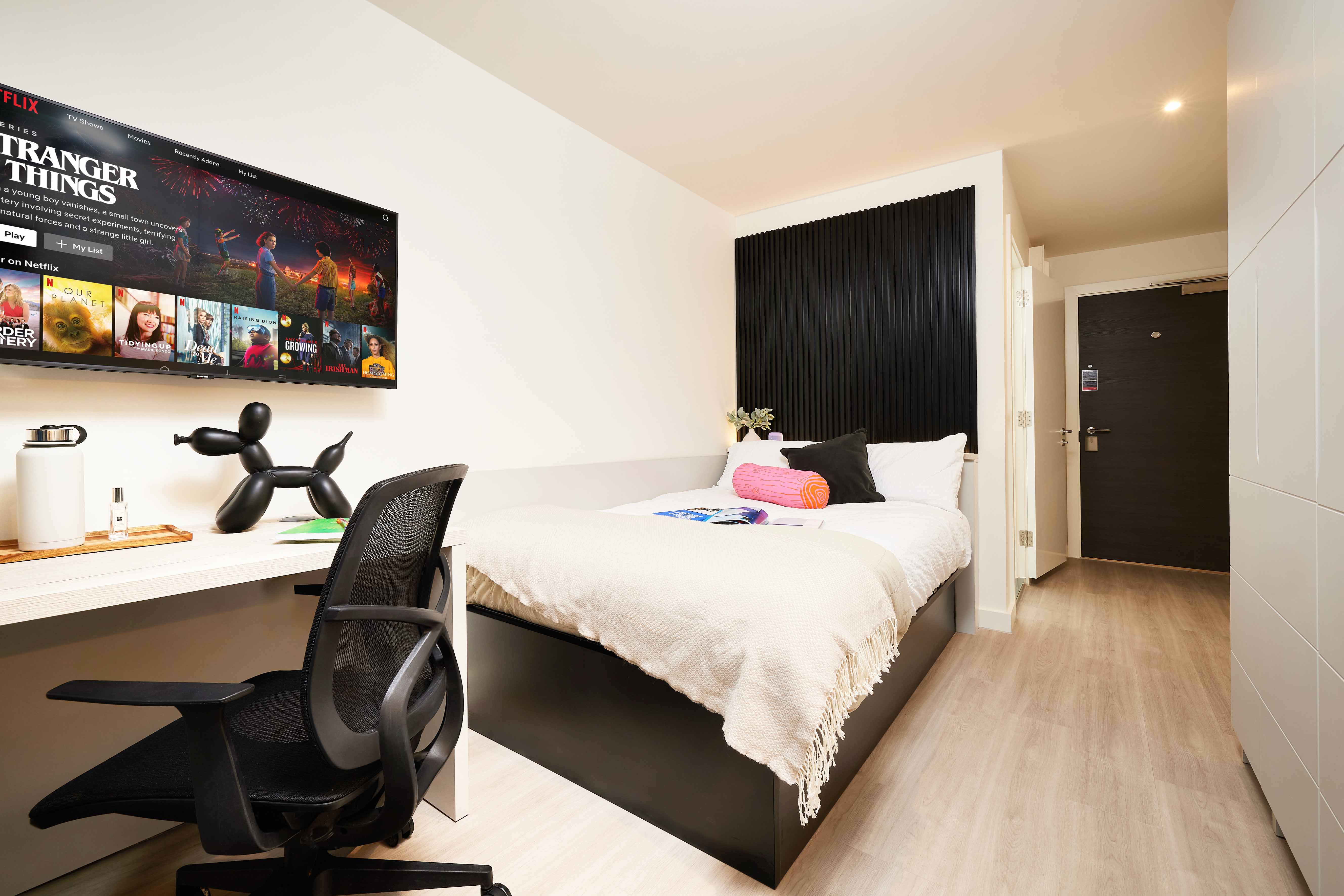 A modern, minimalist dorm room features a bed with white bedding and colorful pillows. A small desk with a black chair faces a wall-mounted TV displaying streaming content. The room has light wood flooring and a sleek, functional design.