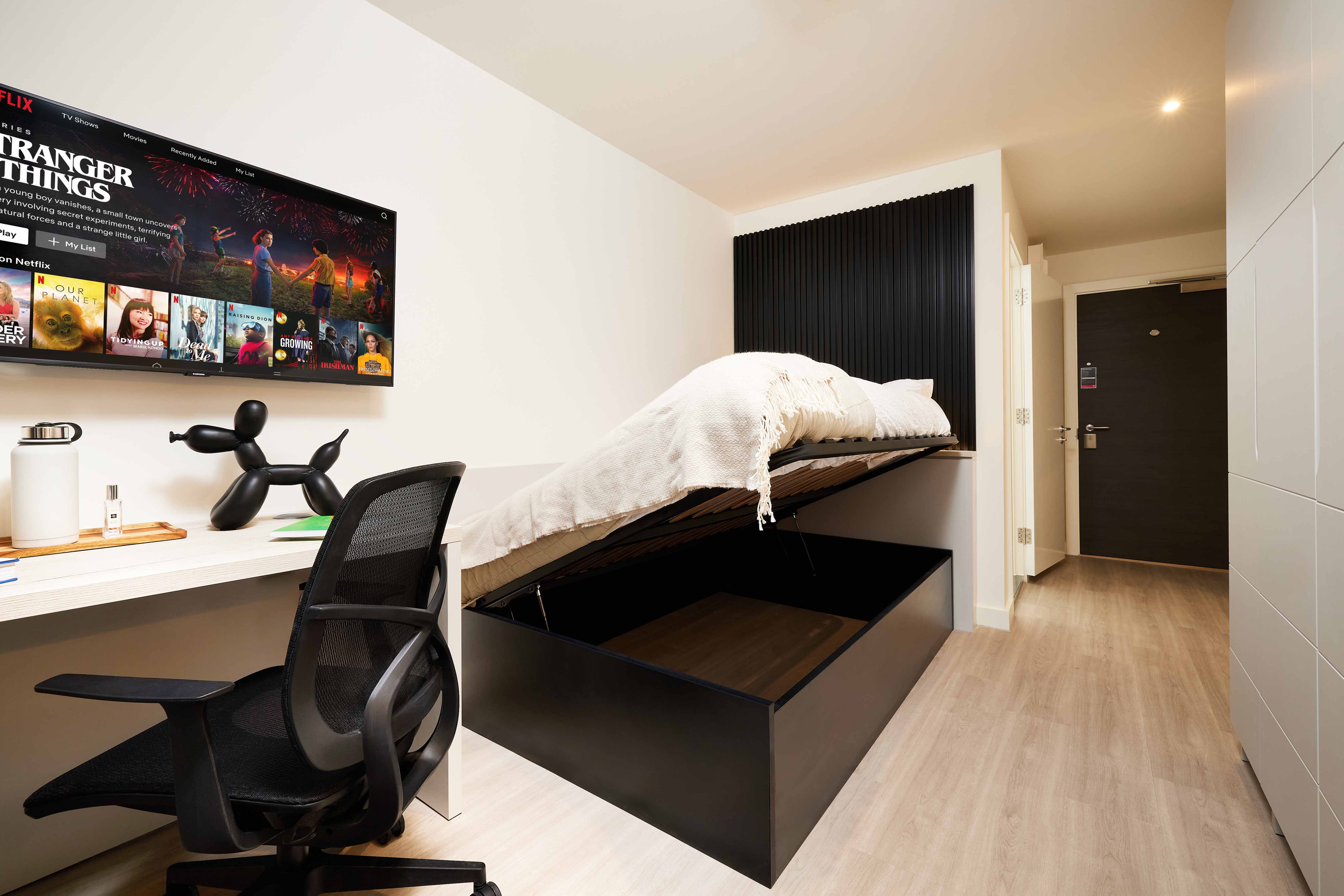 Modern bedroom with a wall-mounted TV displaying Netflix, a pull-down bed partially lifted to reveal storage underneath, sleek desk with chair, and minimalist decor. Bright, functional space with light wooden flooring.