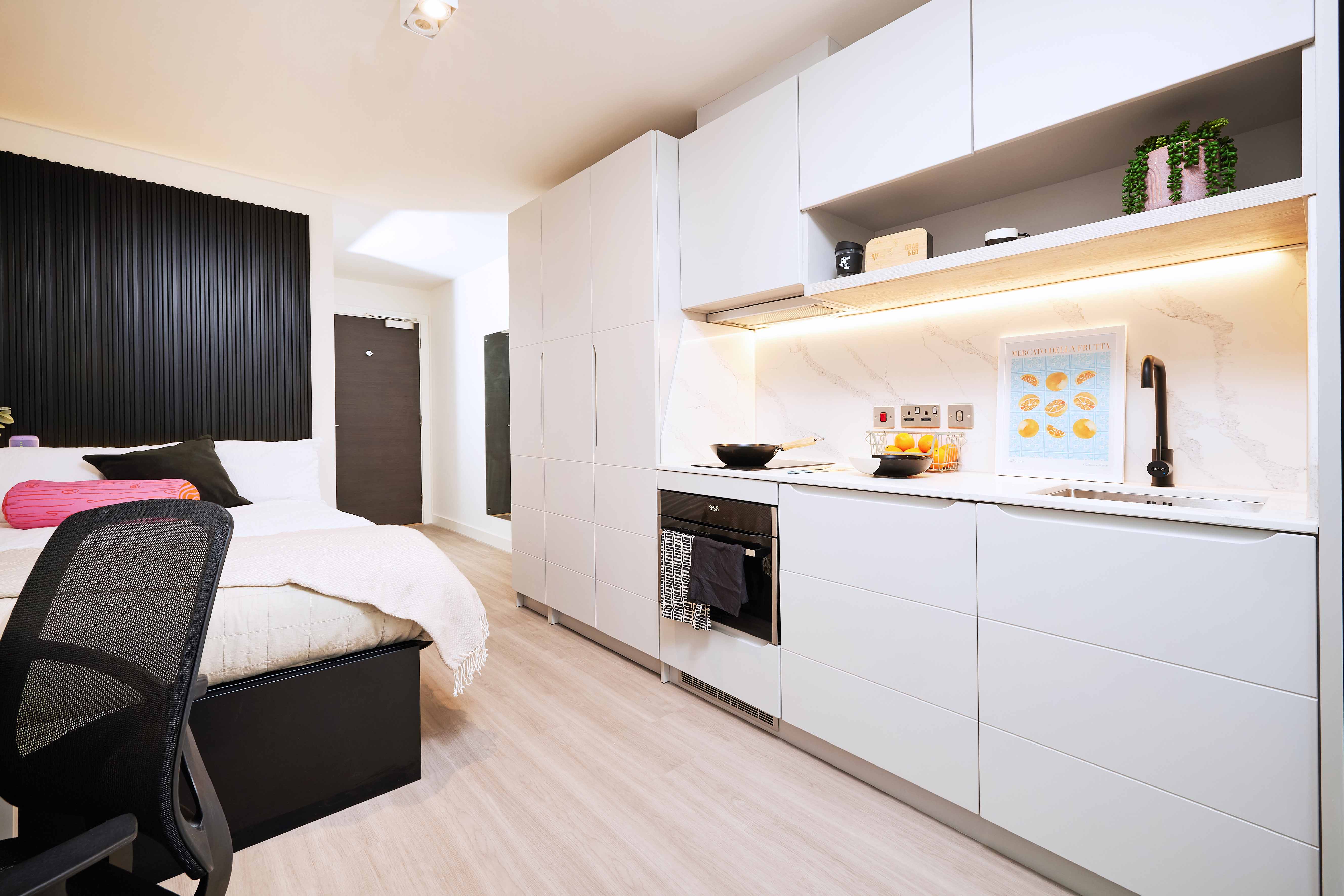 Modern studio apartment with a compact kitchen and cozy bedroom area. The kitchen features white cabinetry, an oven, and sink. The bedroom has a neatly made bed with white linens and a black backboard. A desk chair is positioned beside the bed.