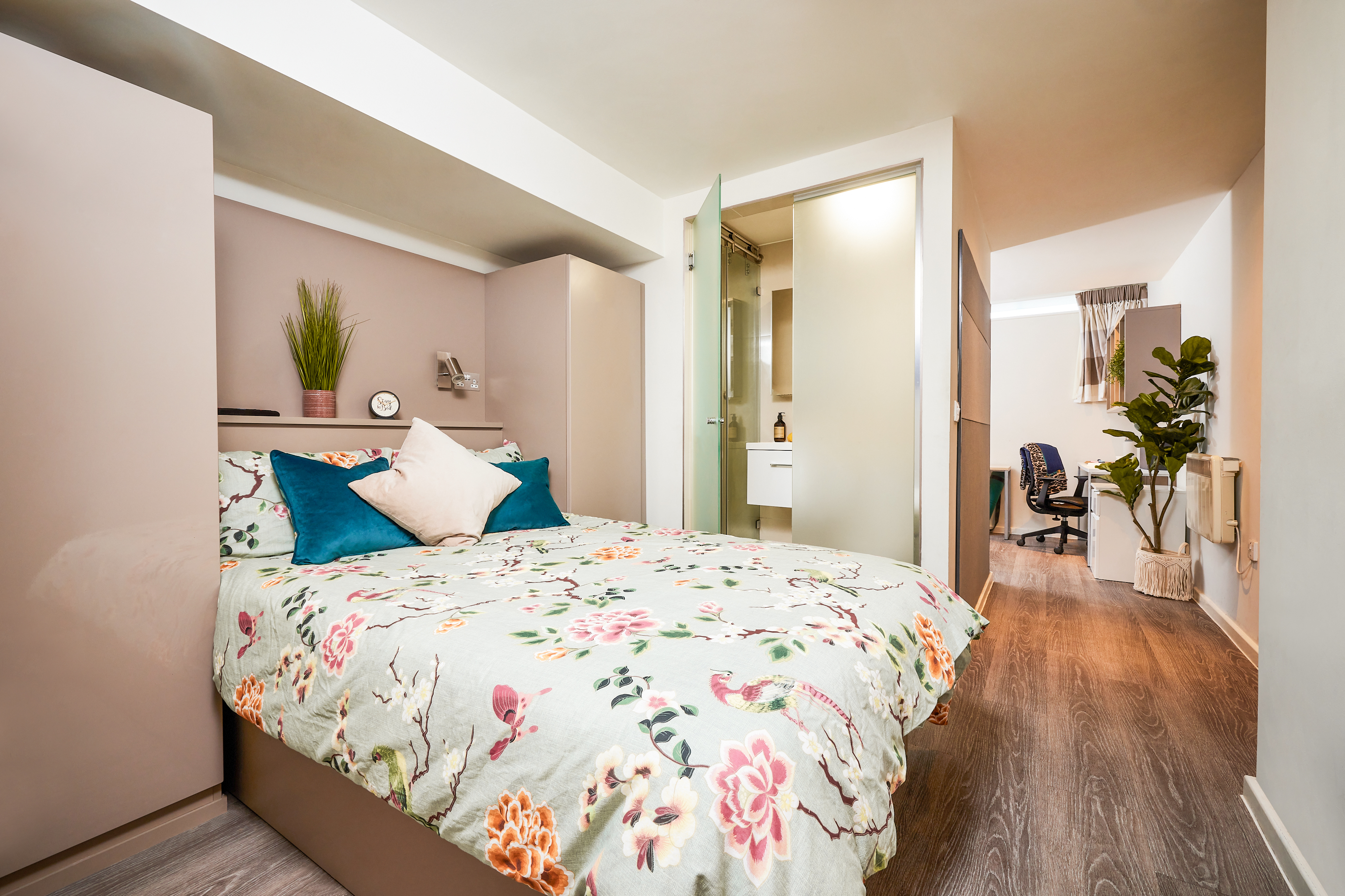 A cozy bedroom in a luxury student accommodation features a floral-patterned bed with colorful pillows, flanked by taupe wardrobes. A bathroom door is partially open, and a study area with a desk and chair is visible. A potted plant adds greenery to the light-wood floor.