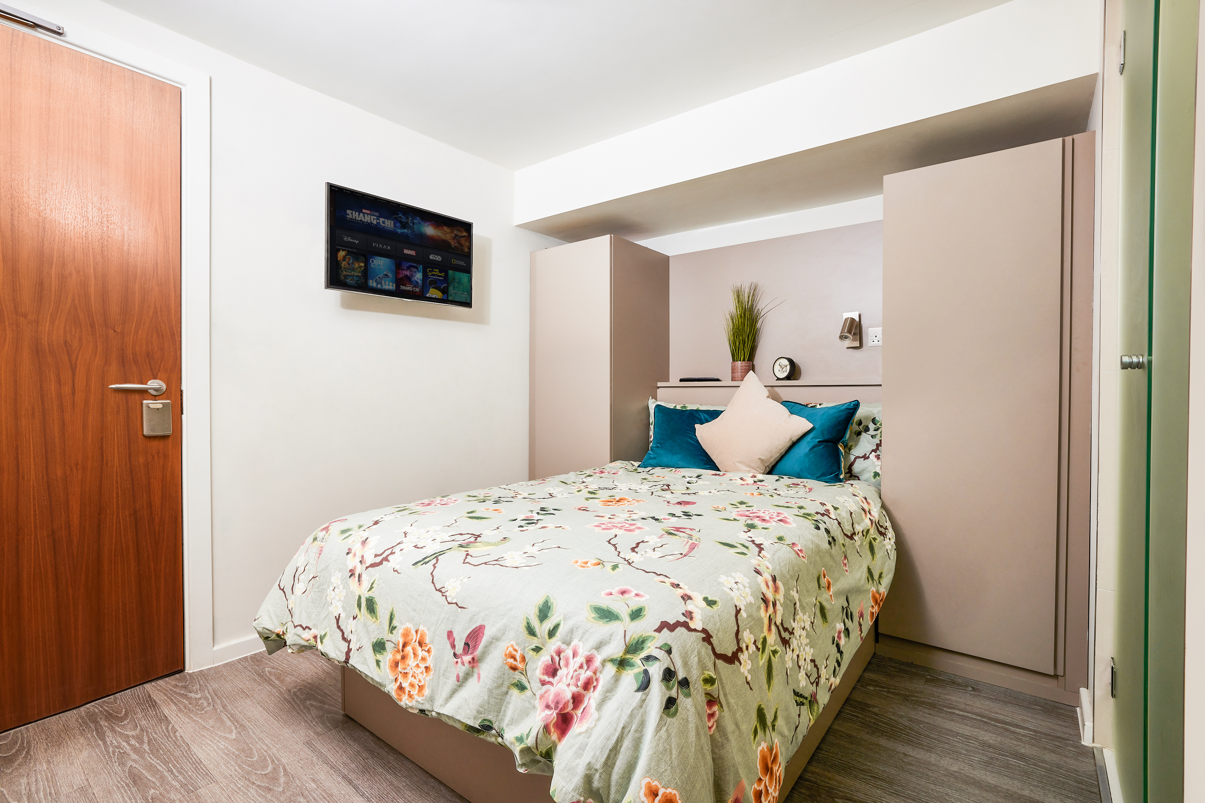 A cozy bedroom in luxury student accommodation features a floral-patterned bedspread on a double bed. Teal and beige pillows rest at the head. A wall-mounted TV is on the left, with light-colored wardrobes flanking the space. A plant and clock sit on a small shelf above the bed.