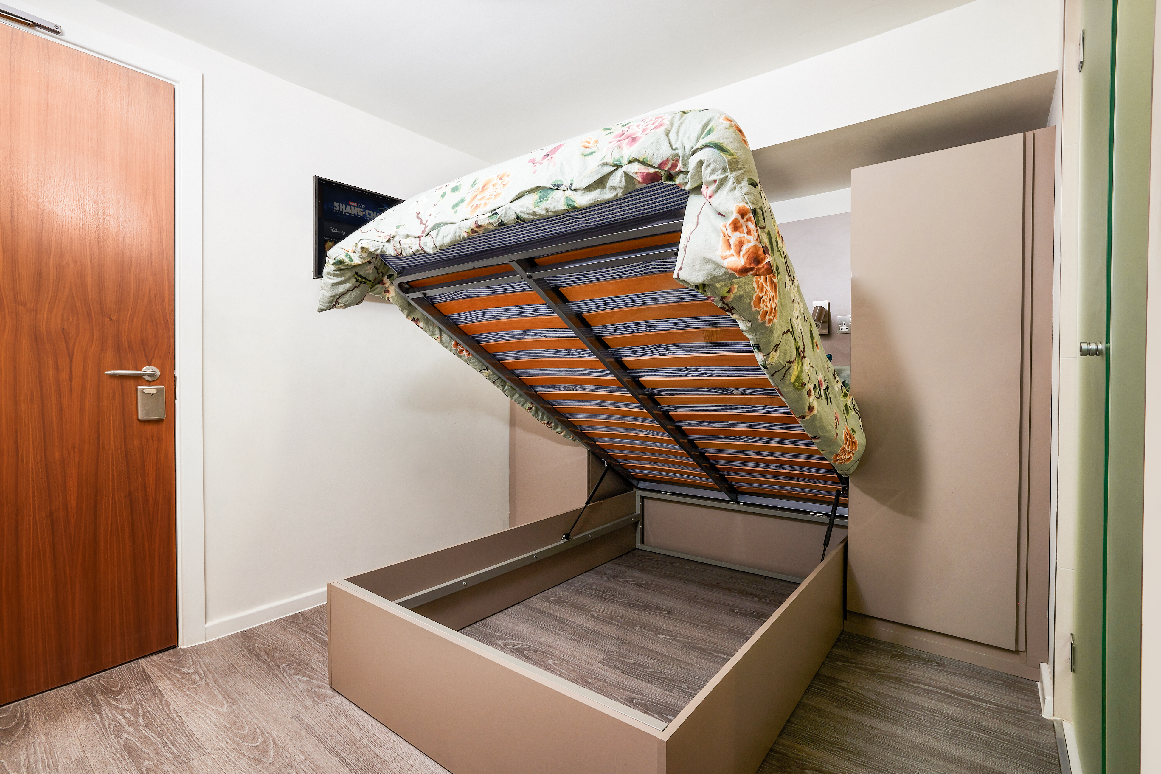 This luxury student accommodation boasts a room with a wooden floor, featuring a bed with its mattress raised for extra storage. The bed is adorned with a floral-patterned cover. To the left, there's a wooden door, while a small flat-screen TV is conveniently mounted on the wall.