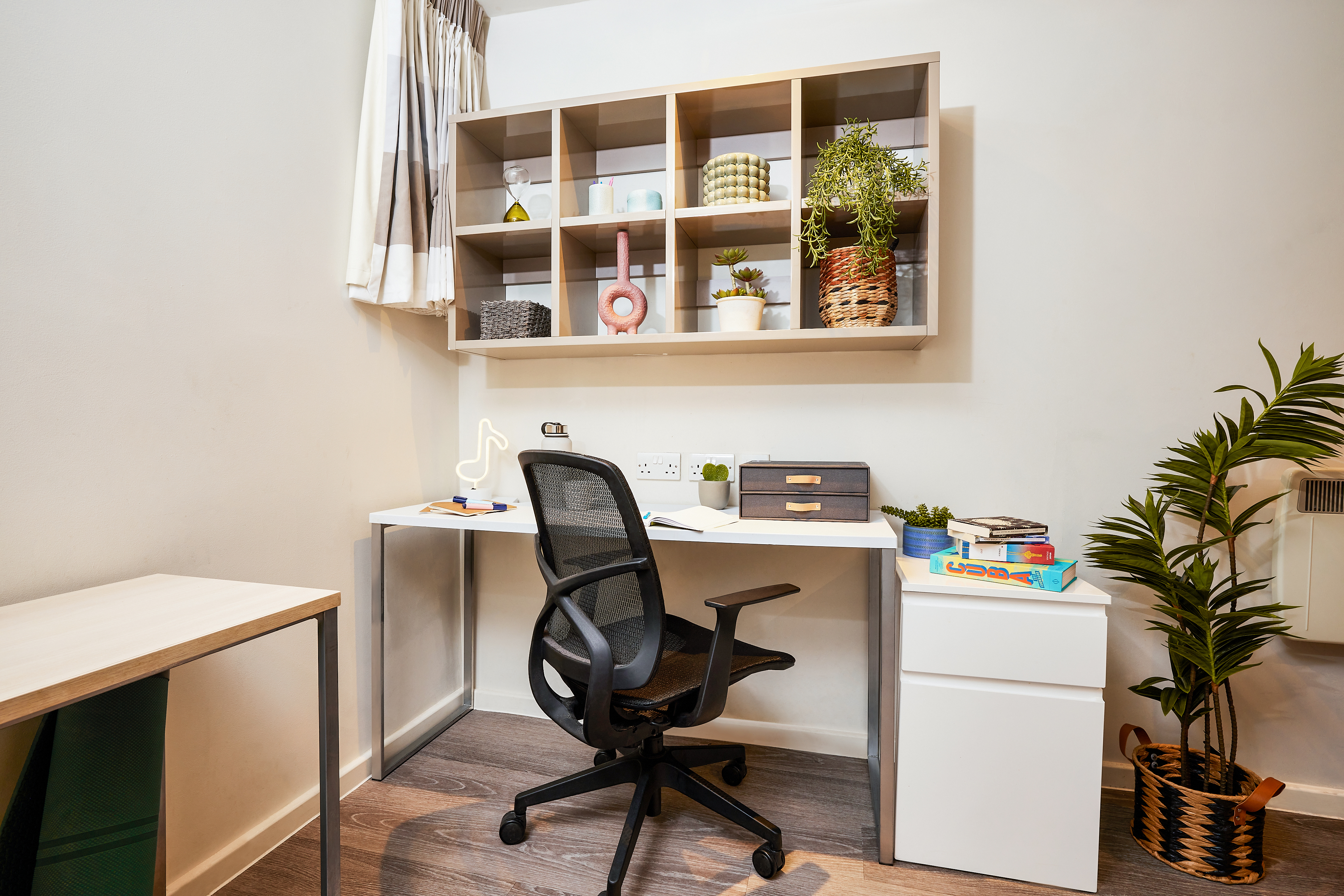 A tidy home office with a modern desk featuring a black office chair, white drawers, and various stationery evokes the feel of luxury student accommodation. Above the desk, a wall-mounted shelving unit holds books, plants, and decorative items. A potted plant rests on the floor nearby.