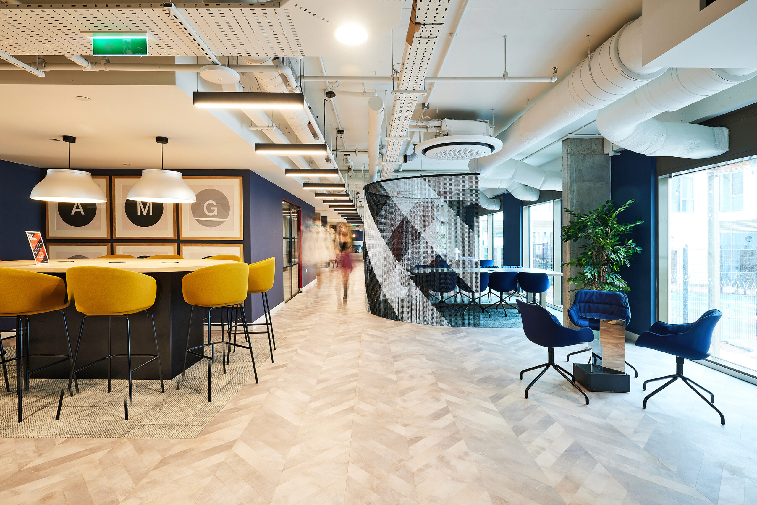 Modern office space, perfect for student accommodation in Coventry, features a large table with yellow chairs on the left and geometric wall art in the center. Blue chairs by tables are near large windows. An exposed ceiling and a mix of wood and carpet flooring complete the contemporary vibe.