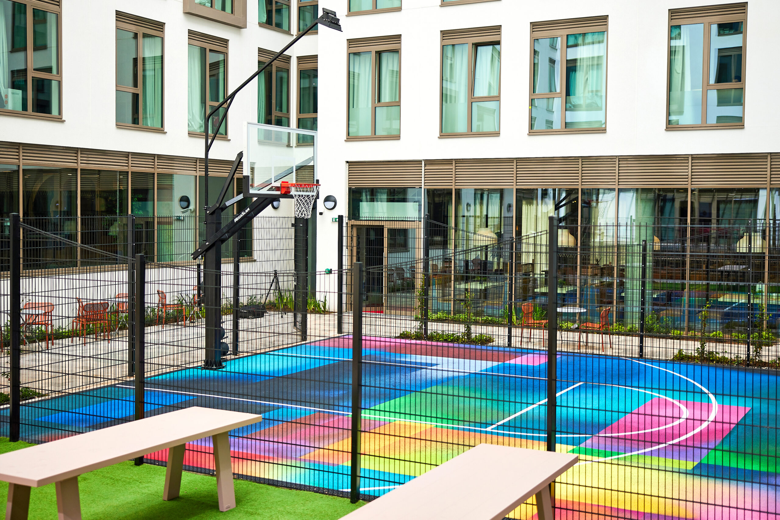 A colorful outdoor basketball court with a geometric pattern is enclosed by a black fence. Located near student accommodation in Coventry, the court sits next to a modern building with large windows. Two beige benches rest on the green turf adjacent to this vibrant recreational space.