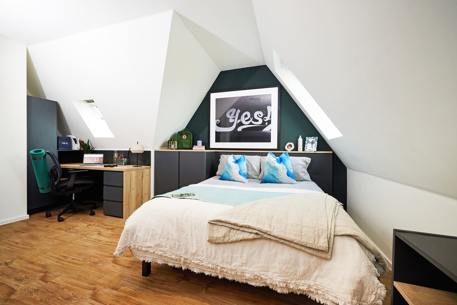 Attic bedroom with a slanted ceiling, featuring a bed with white and blue bedding, a desk with a green chair, and wooden flooring. A large framed print reading "Yes!" is on the wall above the bed. Bright natural light enters through skylights.