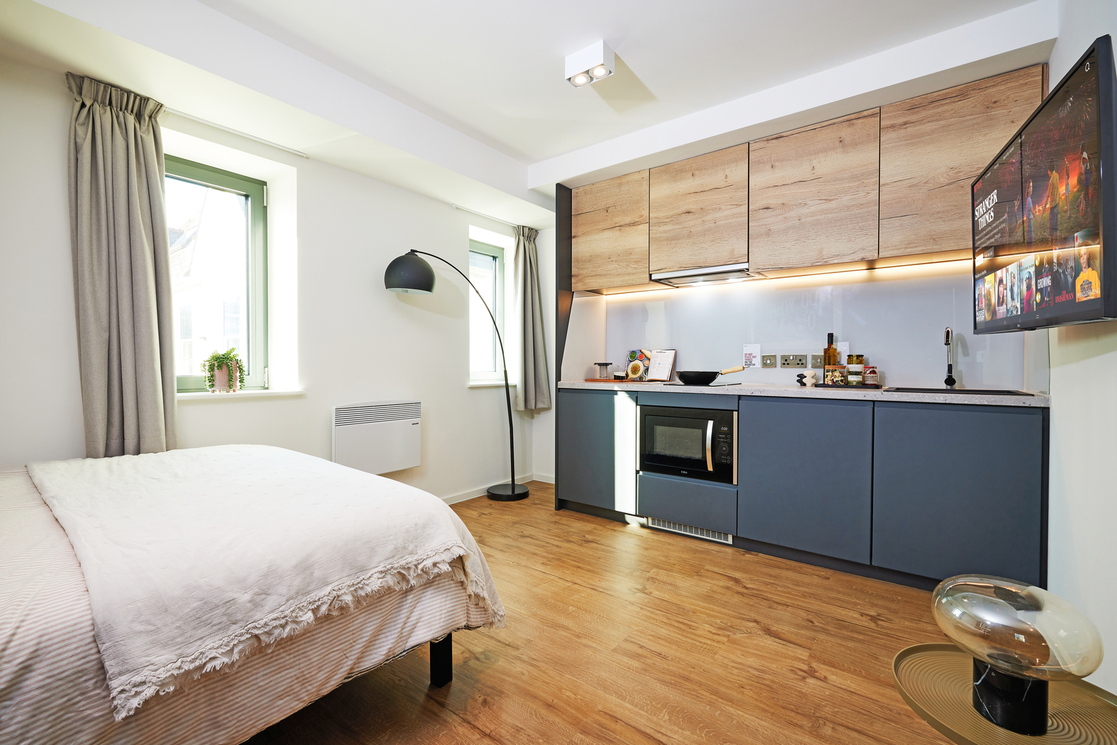 Modern studio apartment with wood flooring, featuring a bed, a kitchenette with blue cabinets and wooden accents, a mounted TV, and a floor lamp. Two windows with beige curtains allow natural light. A small circular table with a glass top sits near the bed.